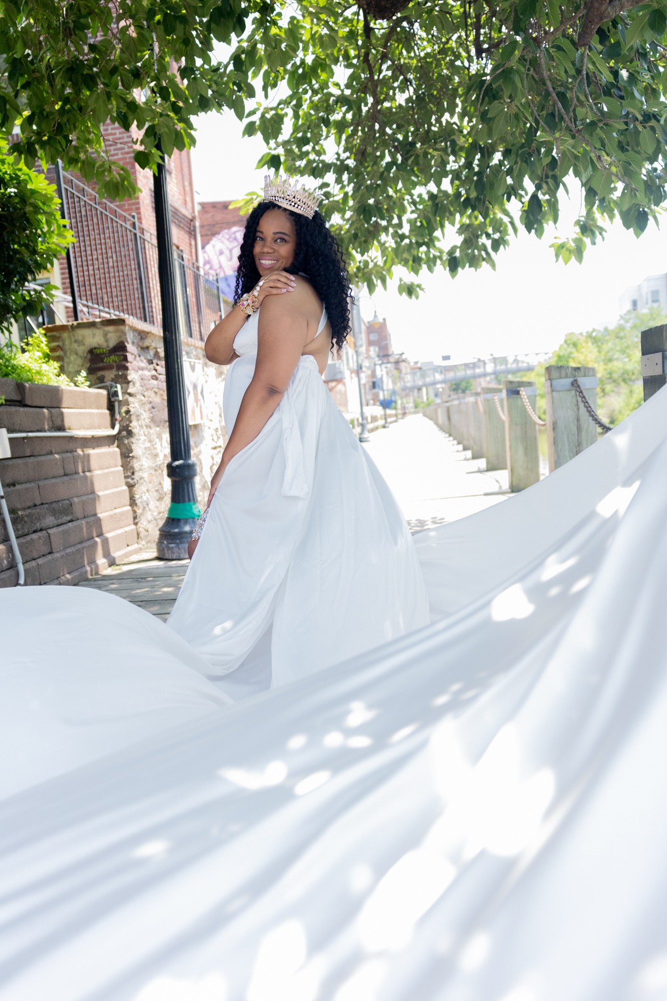 Woman in a white dress standing outdoors under a tree, with a flowing train and a city street background.