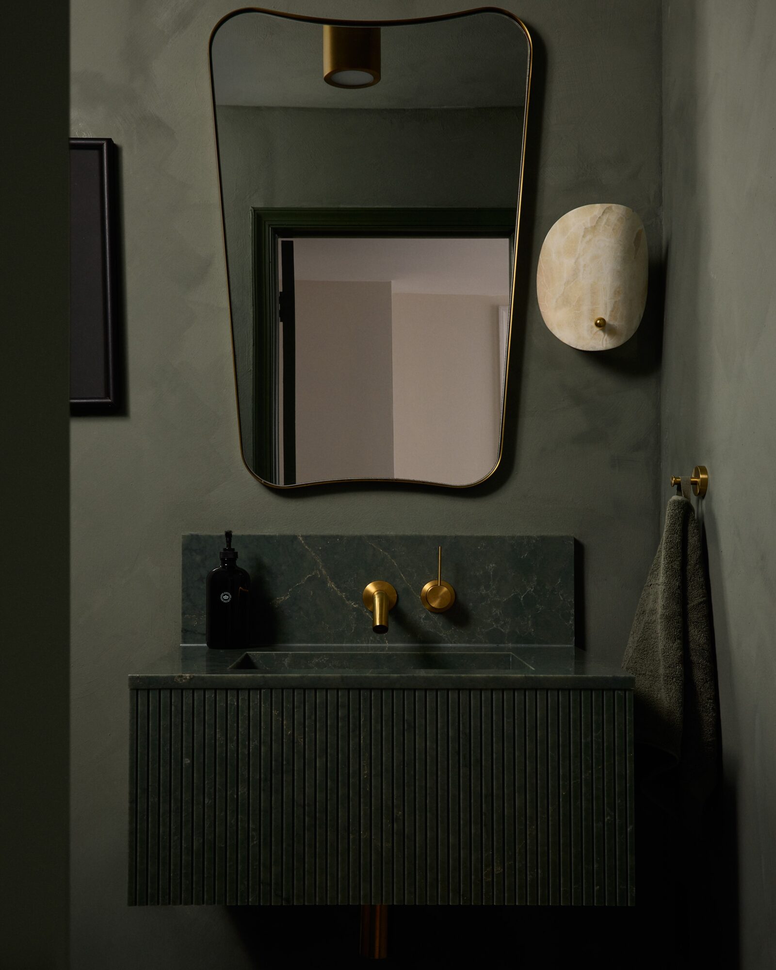 Bathroom with a mirror, sink, and wall-mounted light fixture, all in a modern style with dark and gold accents.