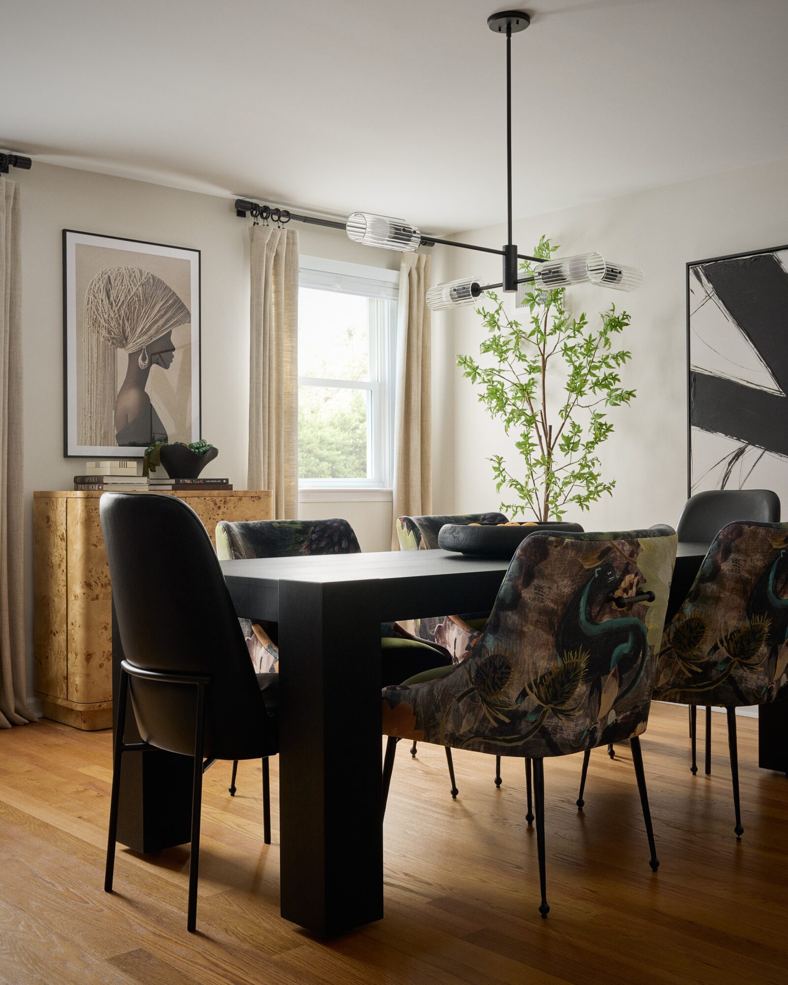 Dining room with black table, six chairs, large window, artwork, and a potted plant, with hardwood floor and neutral decor.
