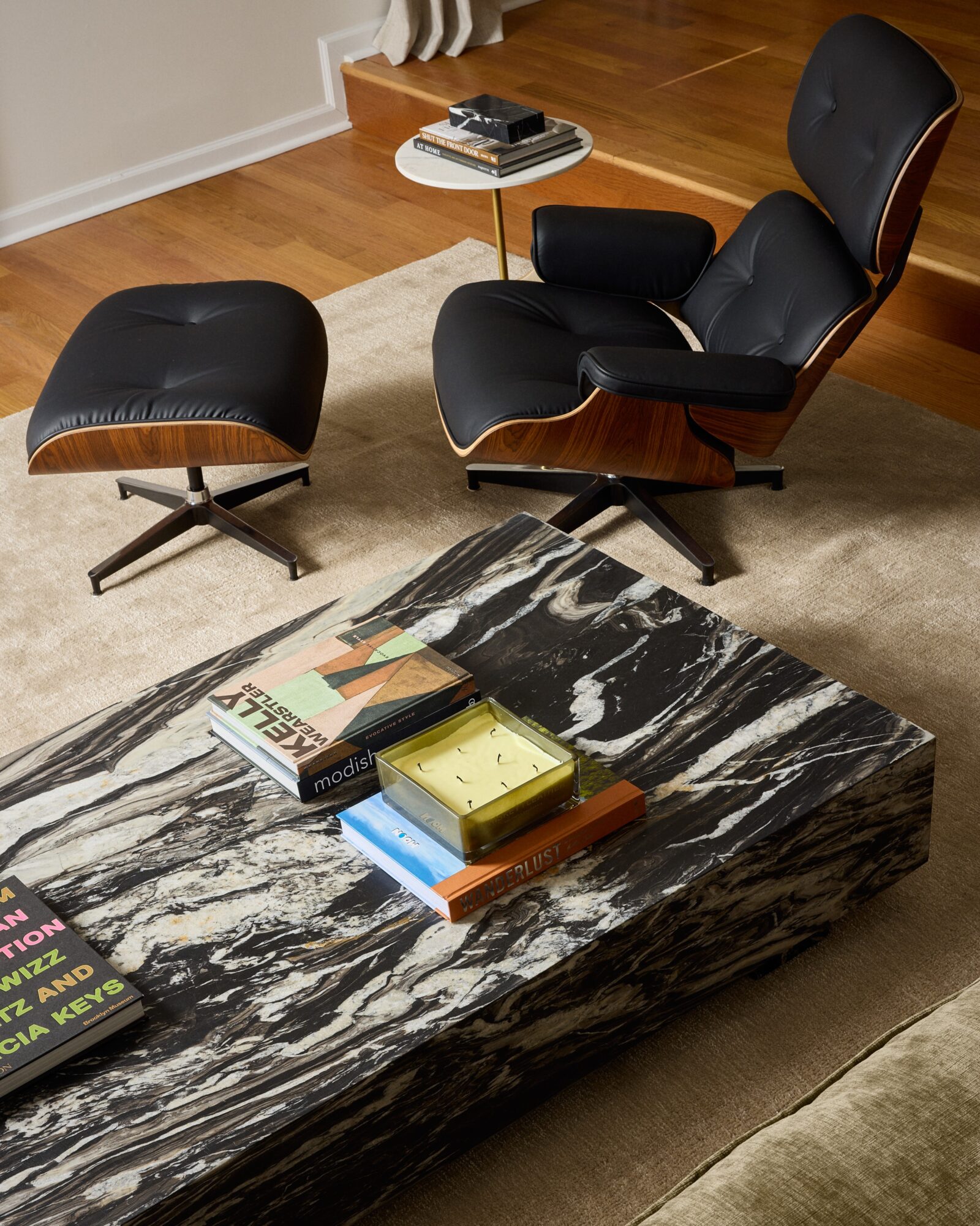 Black lounge chair with matching ottoman, wooden accents, and a small side table with books in a living room. Coffee table with magazines and a yellow box.