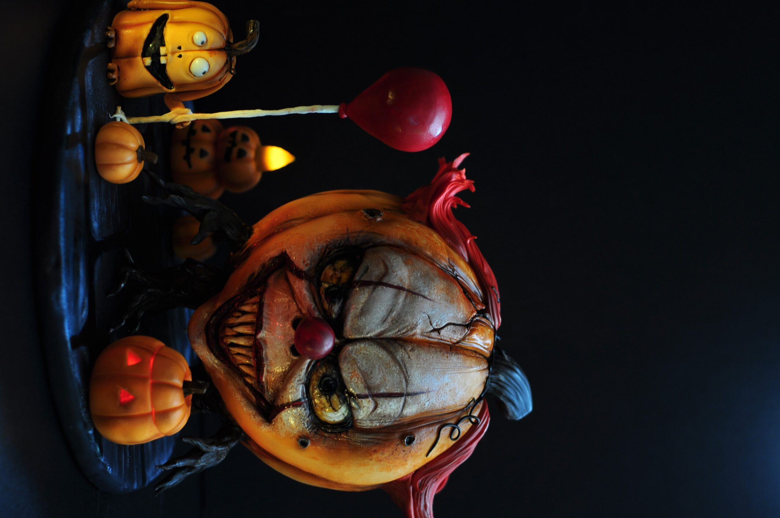 Pumpkin with a carved face, surrounded by small pumpkins and a red balloon, against a dark background.