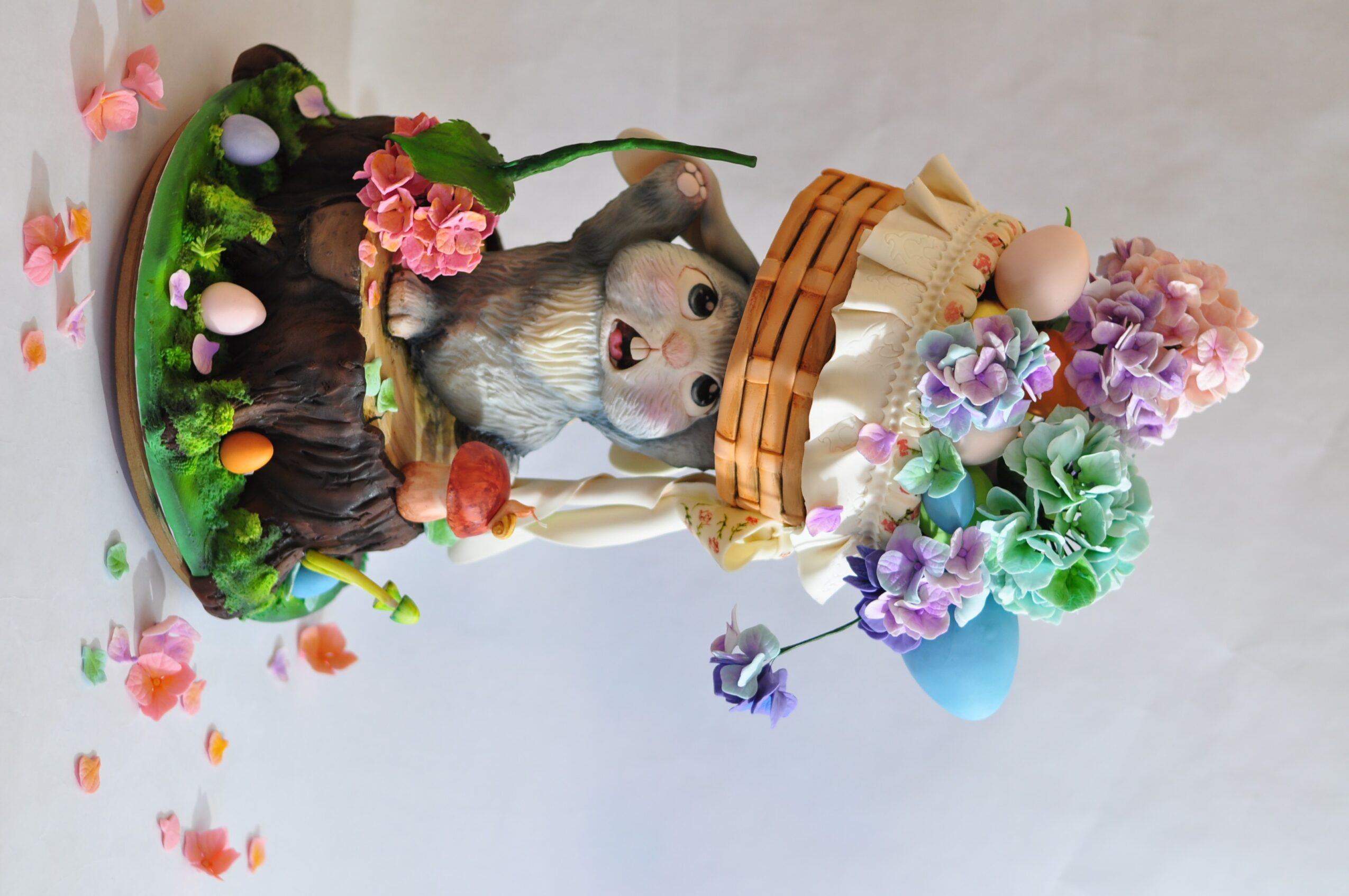 Dog surrounded by colorful flowers and a basket with flowers, with scattered petals on a white surface.