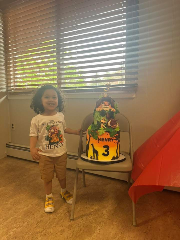 Child with curly hair standing next to a birthday cake with a dinosaur and jungle theme, on a small table.