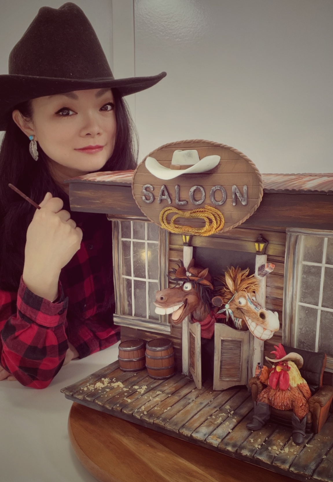 Woman wearing a hat and plaid shirt next to a decorative miniature salon with animals and a sign reading 'Salon'.