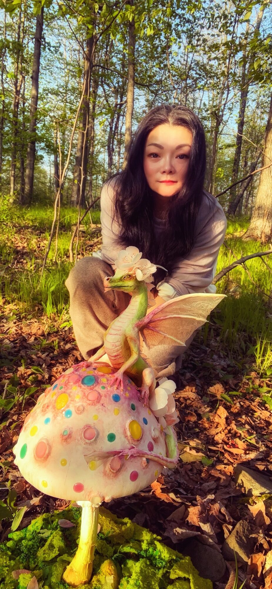 Young woman crouching in a forest with a large, colorful mushroom prop, surrounded by trees and sunlight filtering through leaves.