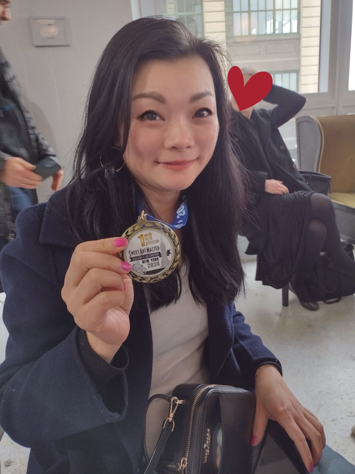 Young woman with dark hair holding a medal, smiling, in an indoor setting with windows and people in the background.