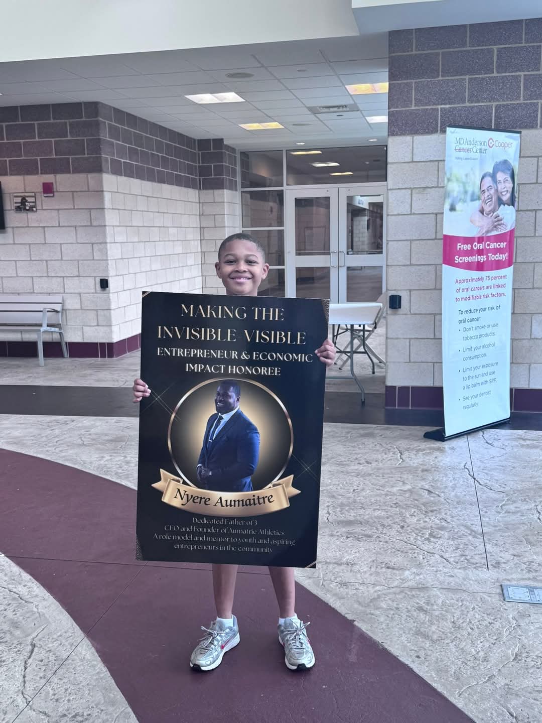 Child holding a large poster board in a building lobby, smiling, with tiled walls and a standing banner nearby.