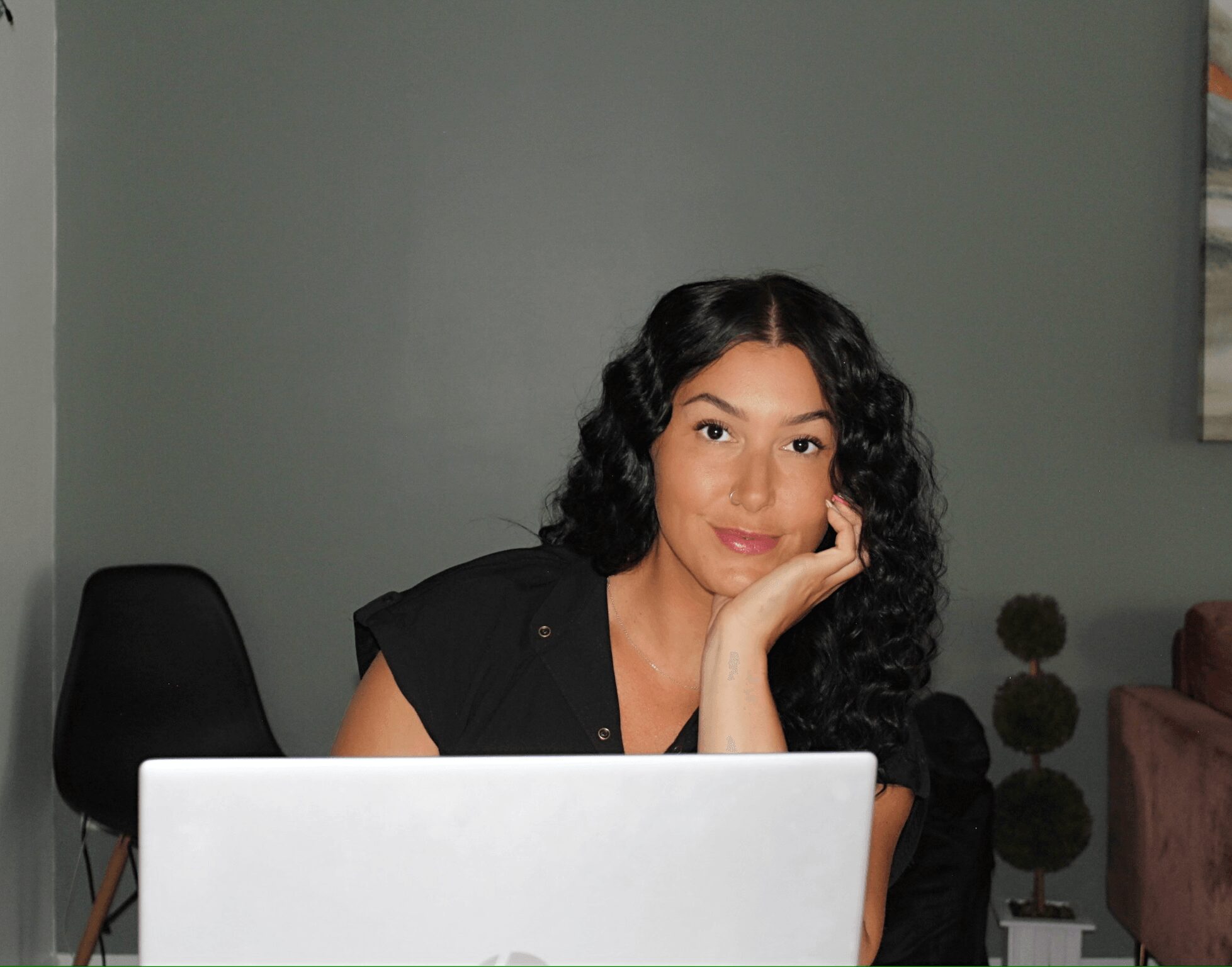 Woman with curly hair sitting at a desk with a laptop, resting her face on her hand, in a room with green walls.
