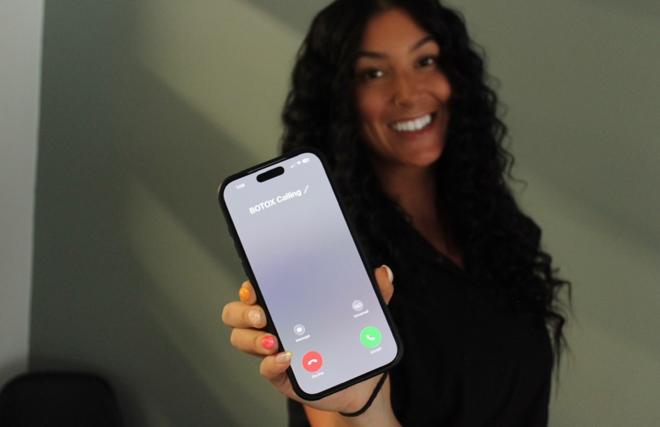 Woman with curly hair smiling and holding a phone showing an incoming call, standing against a green wall.