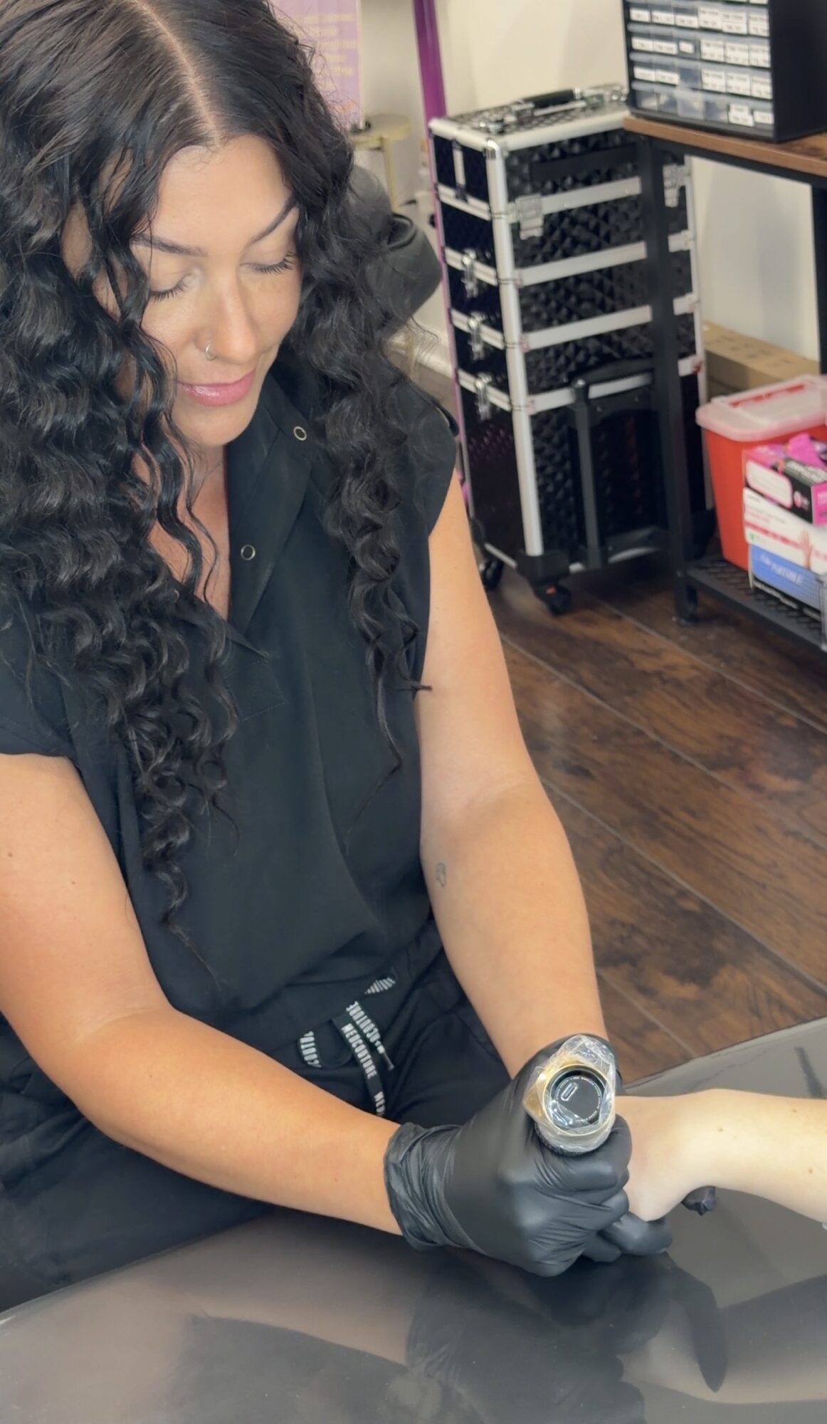 Woman with curly hair wearing black gloves, holding a device, in a room with equipment and shelves.