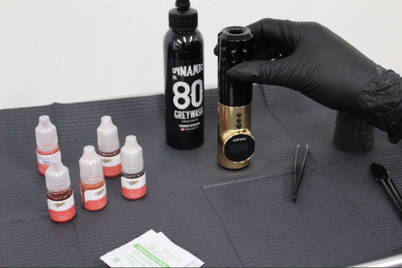Person wearing black glove handling a small black and gold device on a black mat with bottles and tools nearby.