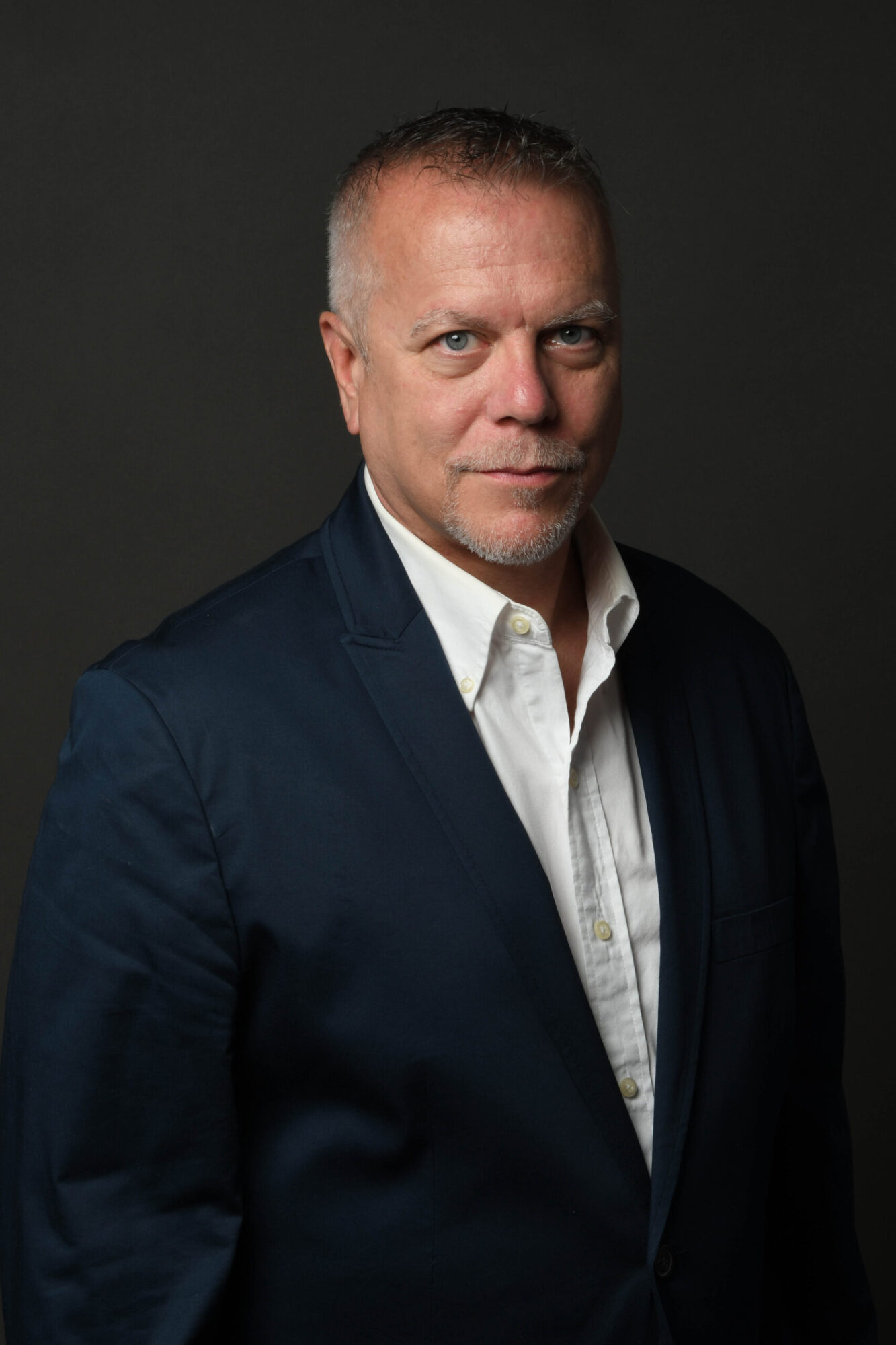 Man in a dark suit and white shirt looking at the camera against a dark background.
