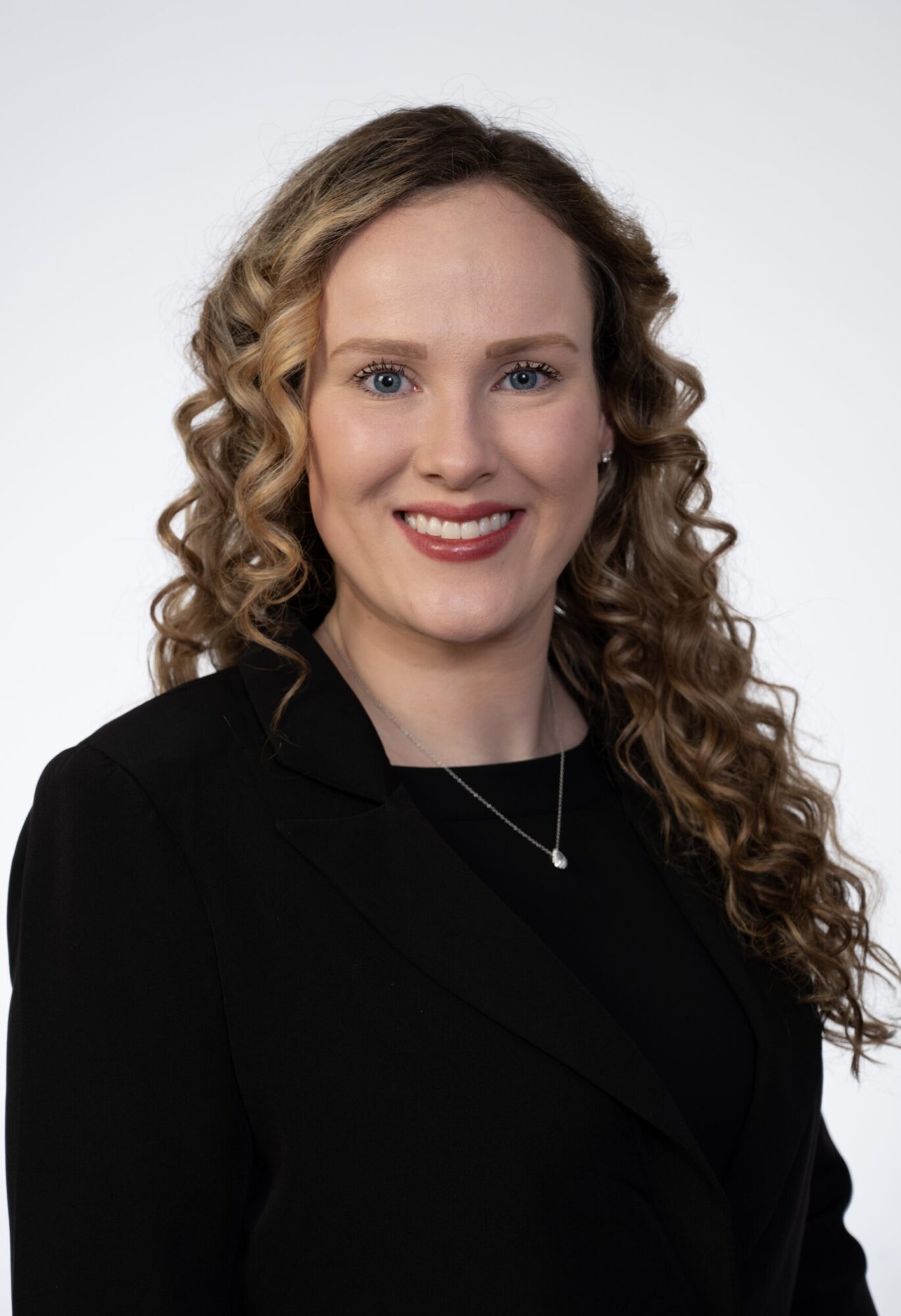 Portrait of a woman with curly hair wearing a black blazer and a necklace, smiling against a plain background.