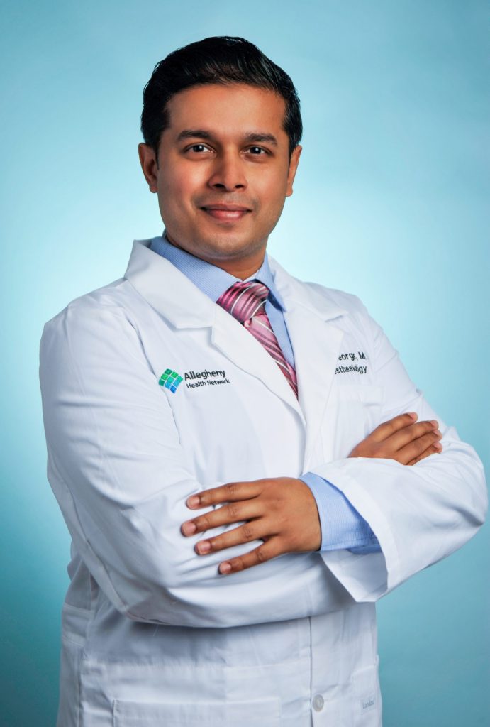 Male doctor in white coat with arms crossed, smiling, against light blue background.