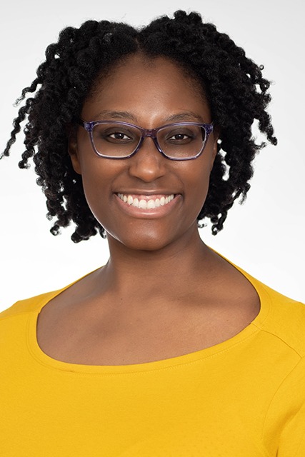 Smiling woman with glasses and curly hair wearing a yellow top, facing forward, against a plain background.