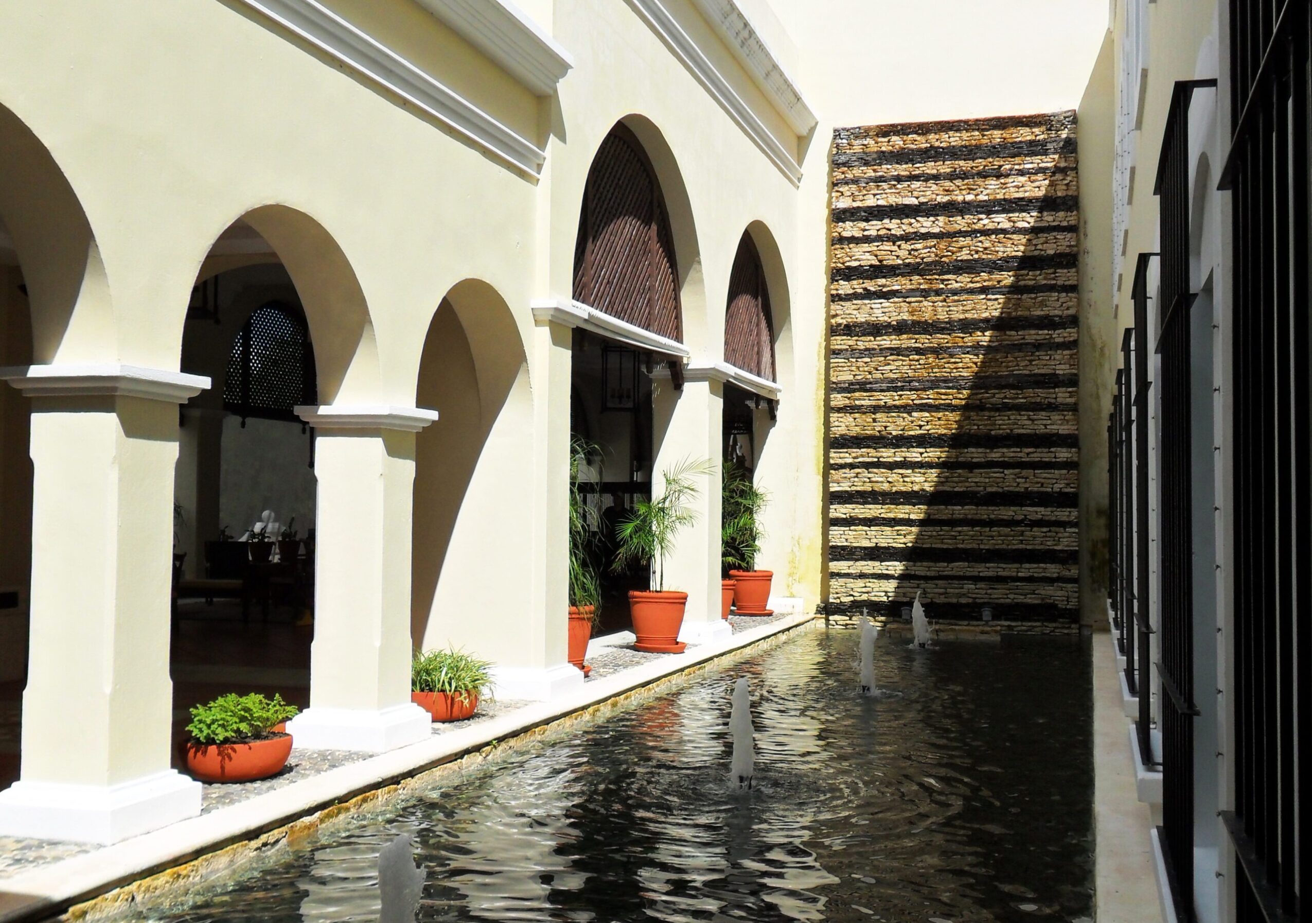 Narrow alley with water feature, arched windows, potted plants, and a staircase against a yellow building.