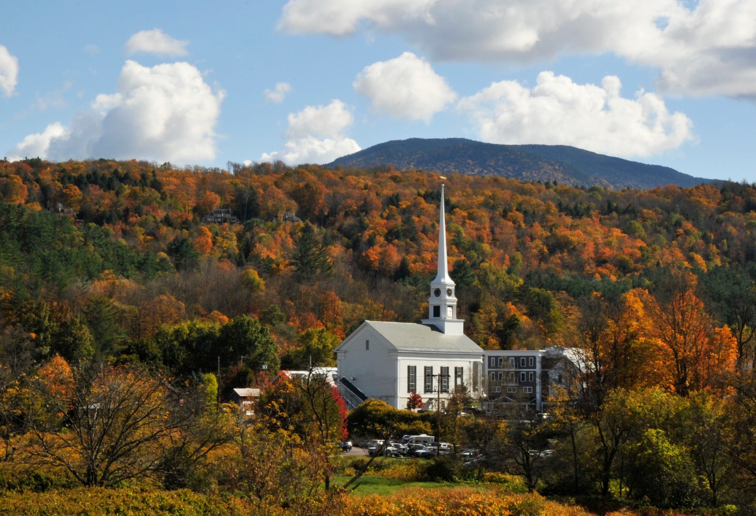 Church with tall steeple surrounded by colorful autumn trees and hills in the background.