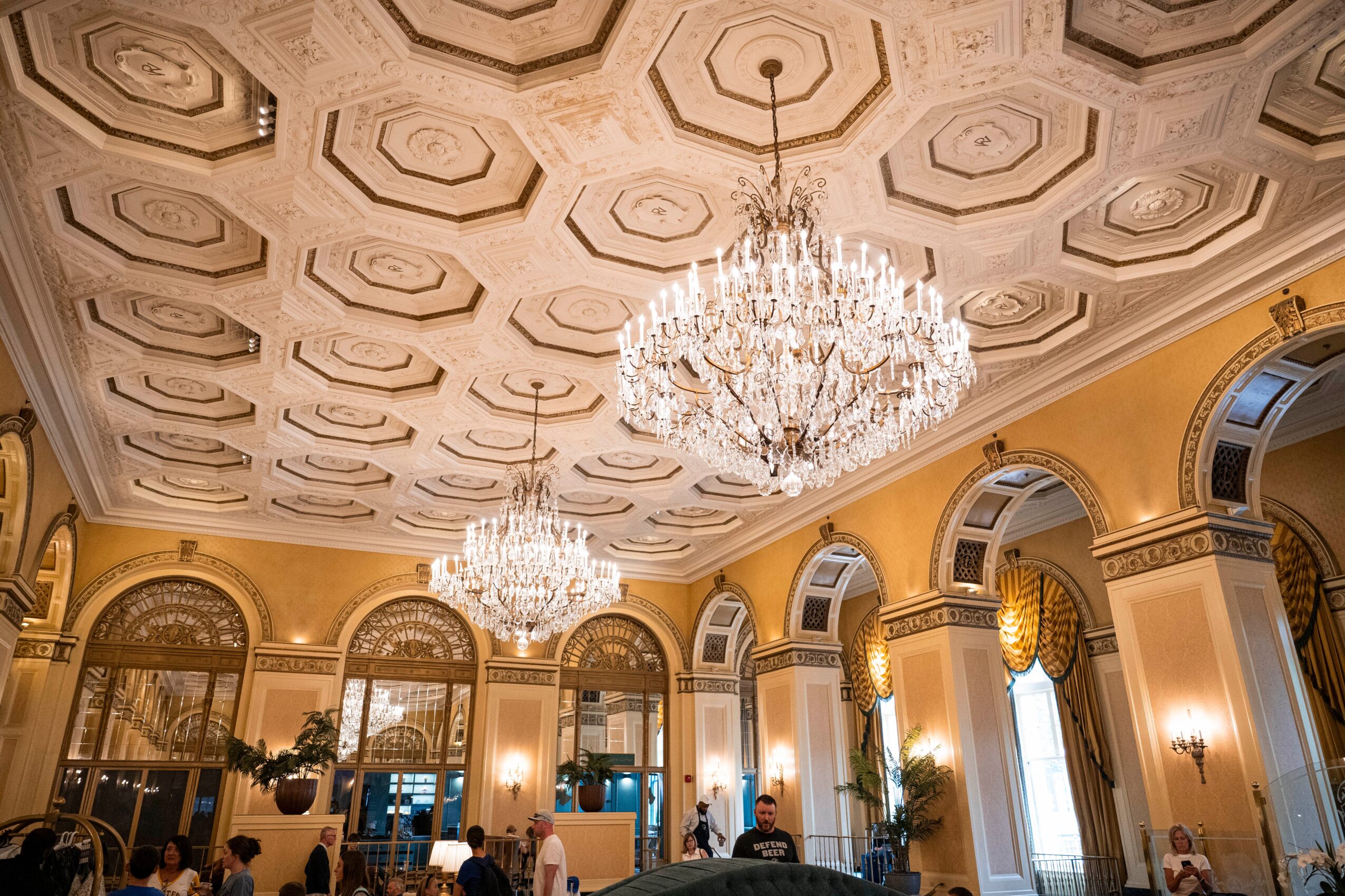 Elegant room with ornate ceiling, large chandeliers, tall windows, and classical decor, filled with people and plants.