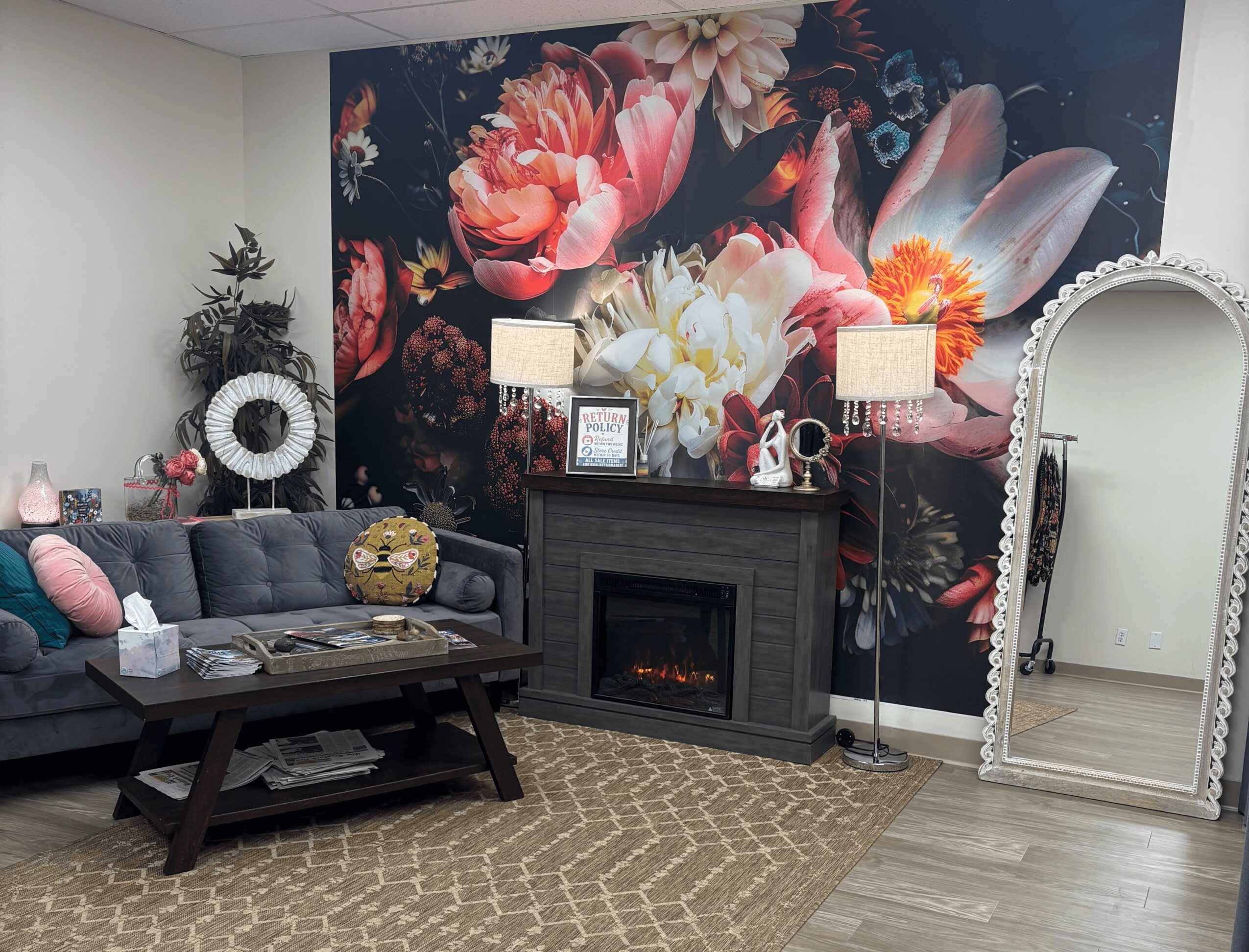 Living room with floral wall art, gray sofa, coffee table, and a large mirror, decorated with plants and lamps.