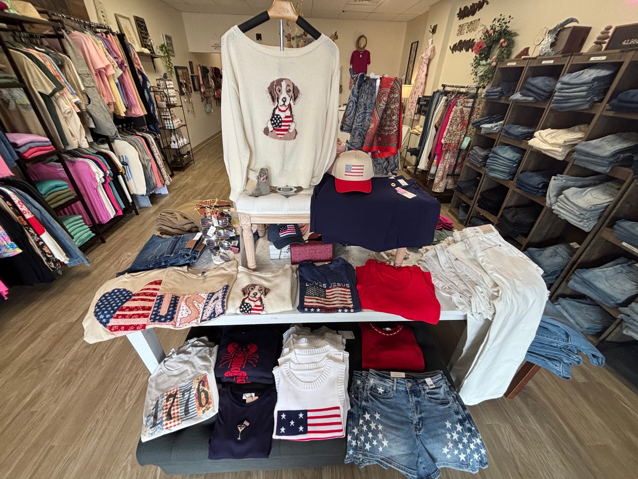 Clothing store display with sweaters, jeans, and accessories on tables and shelves, racks of clothes in background.