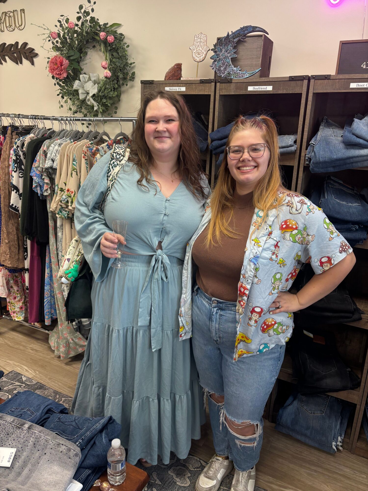 Two women standing in a clothing store, smiling, with shelves of clothes and a floral wreath in the background.