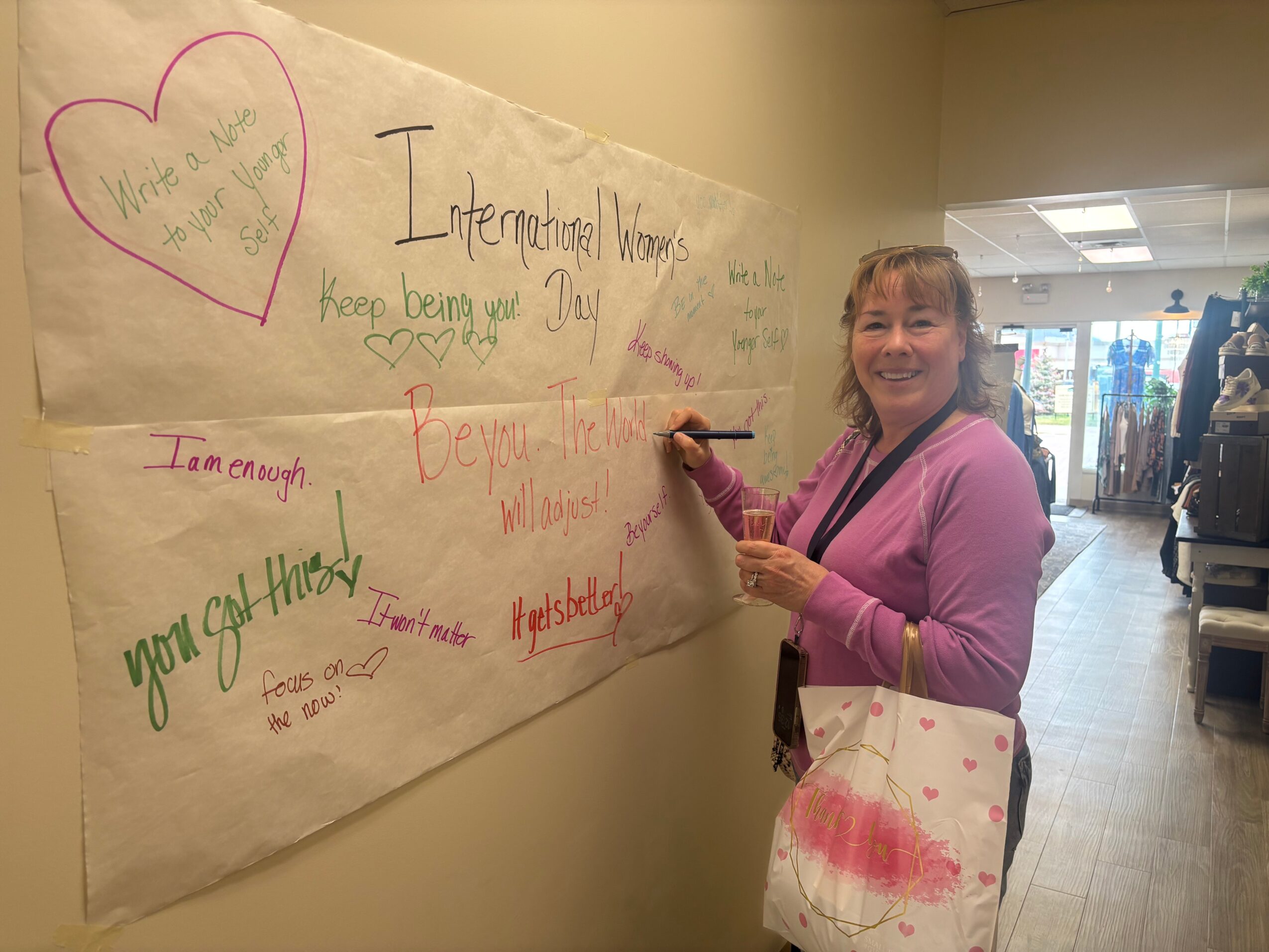Smiling woman in pink sweater holding a marker next to a large whiteboard with handwritten messages, in a well-lit indoor space.