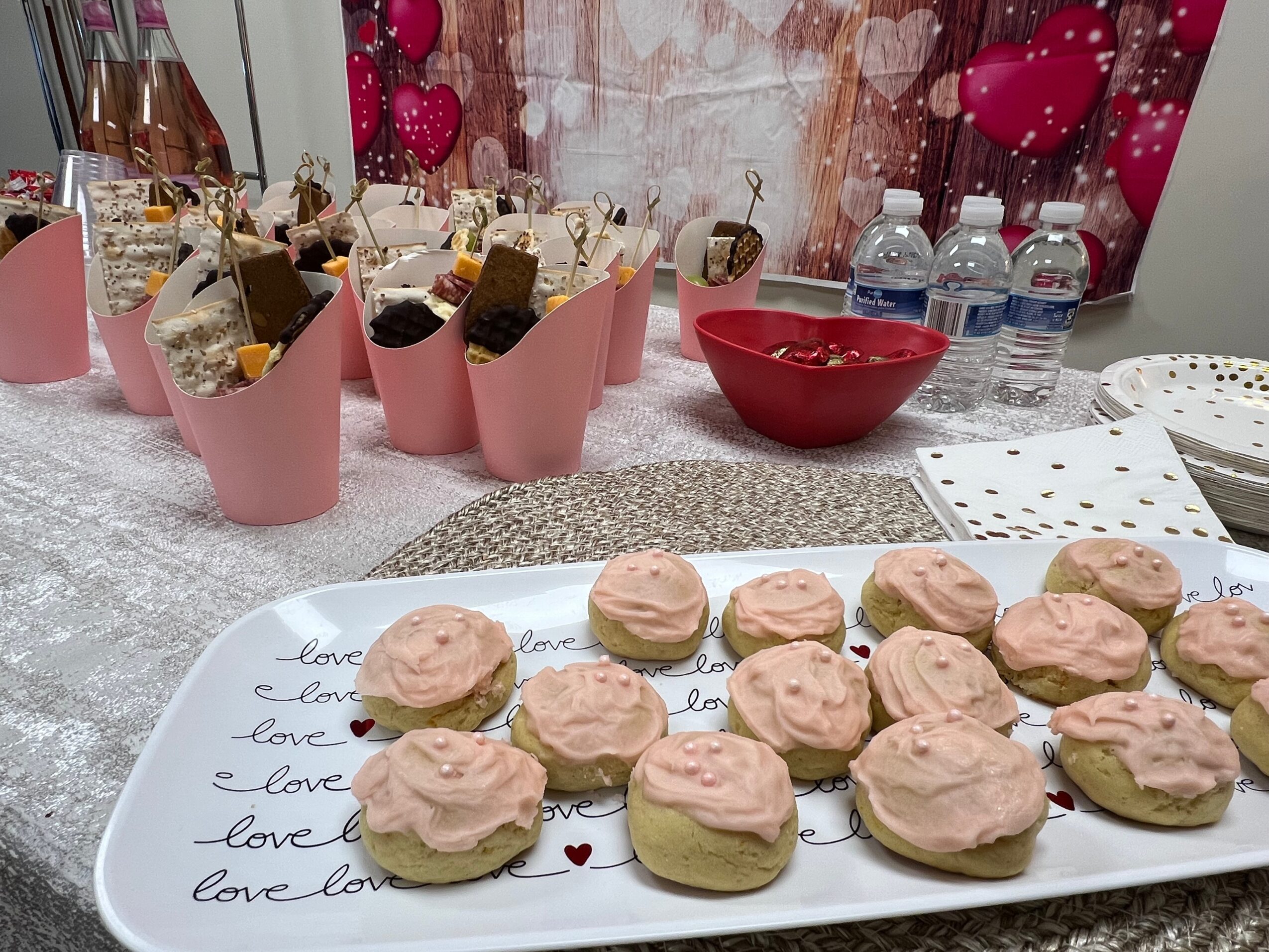 Plate with pink frosted cookies on a table decorated with pink balloons and water bottles, background with pink and white decorations.