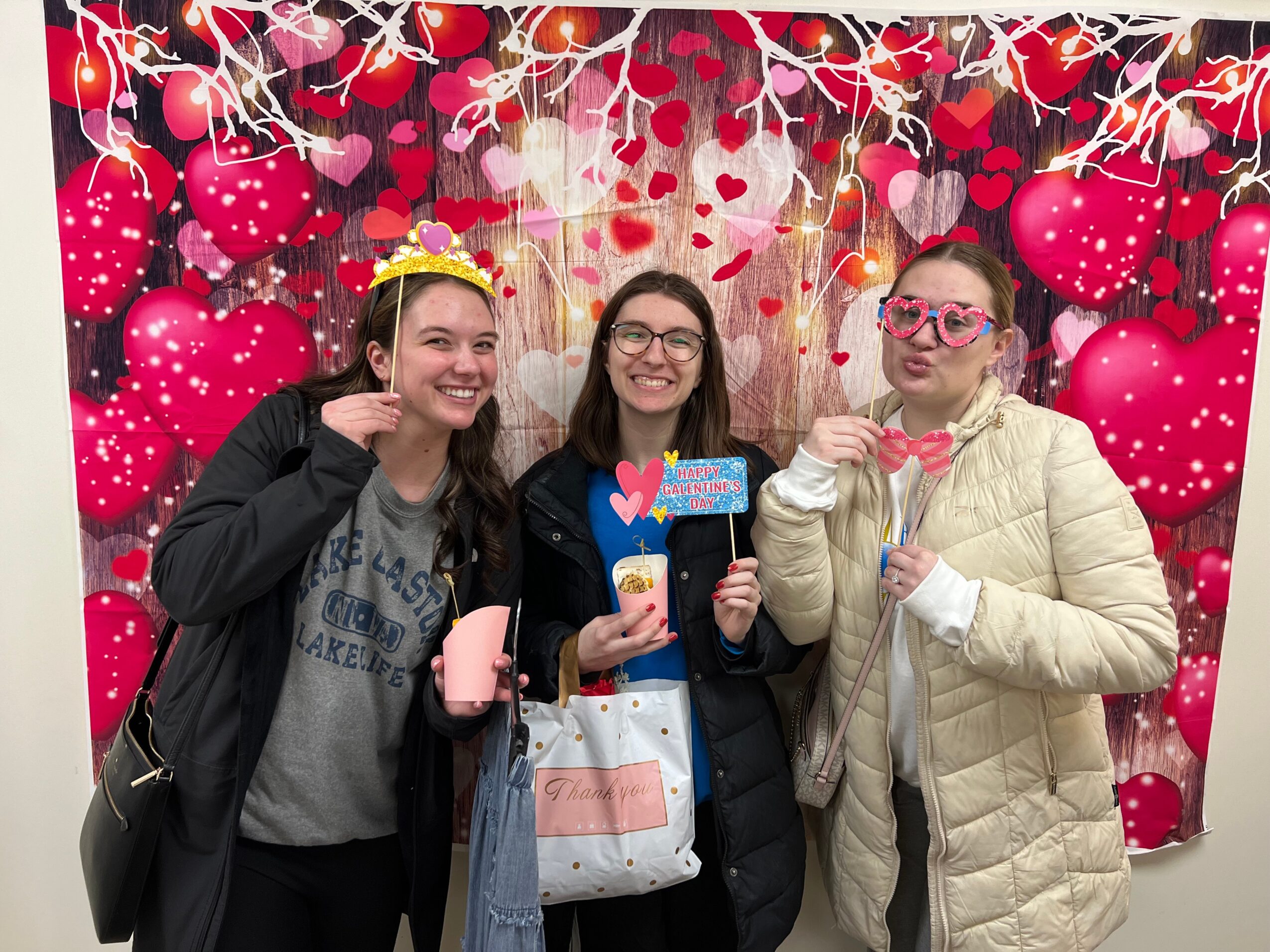 Three women smiling and posing in front of a heart-themed backdrop, holding props, with one wearing glasses and a crown.