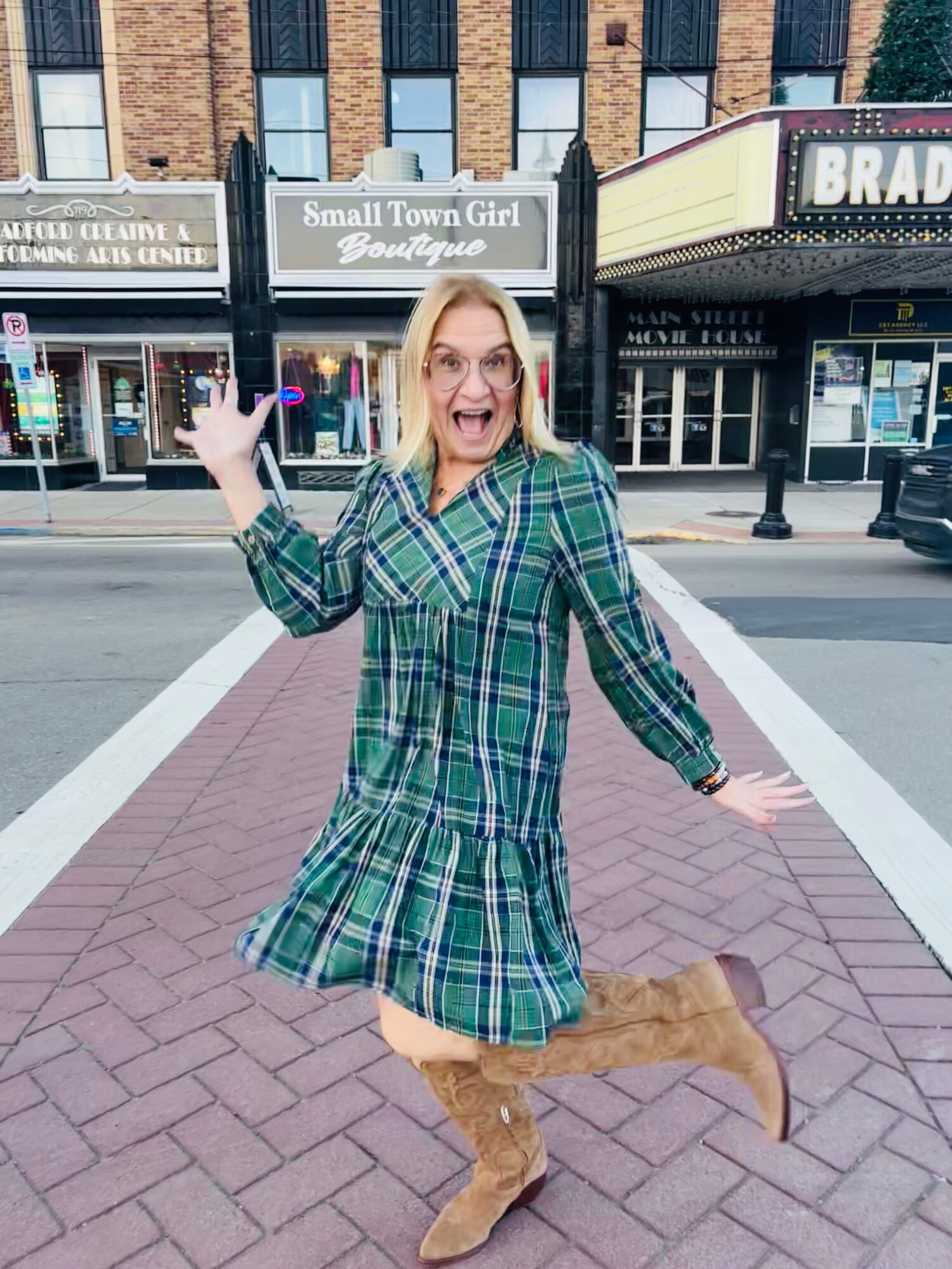 Woman in green plaid dress and tan boots smiling and waving on city street, crossing pedestrian crosswalk.