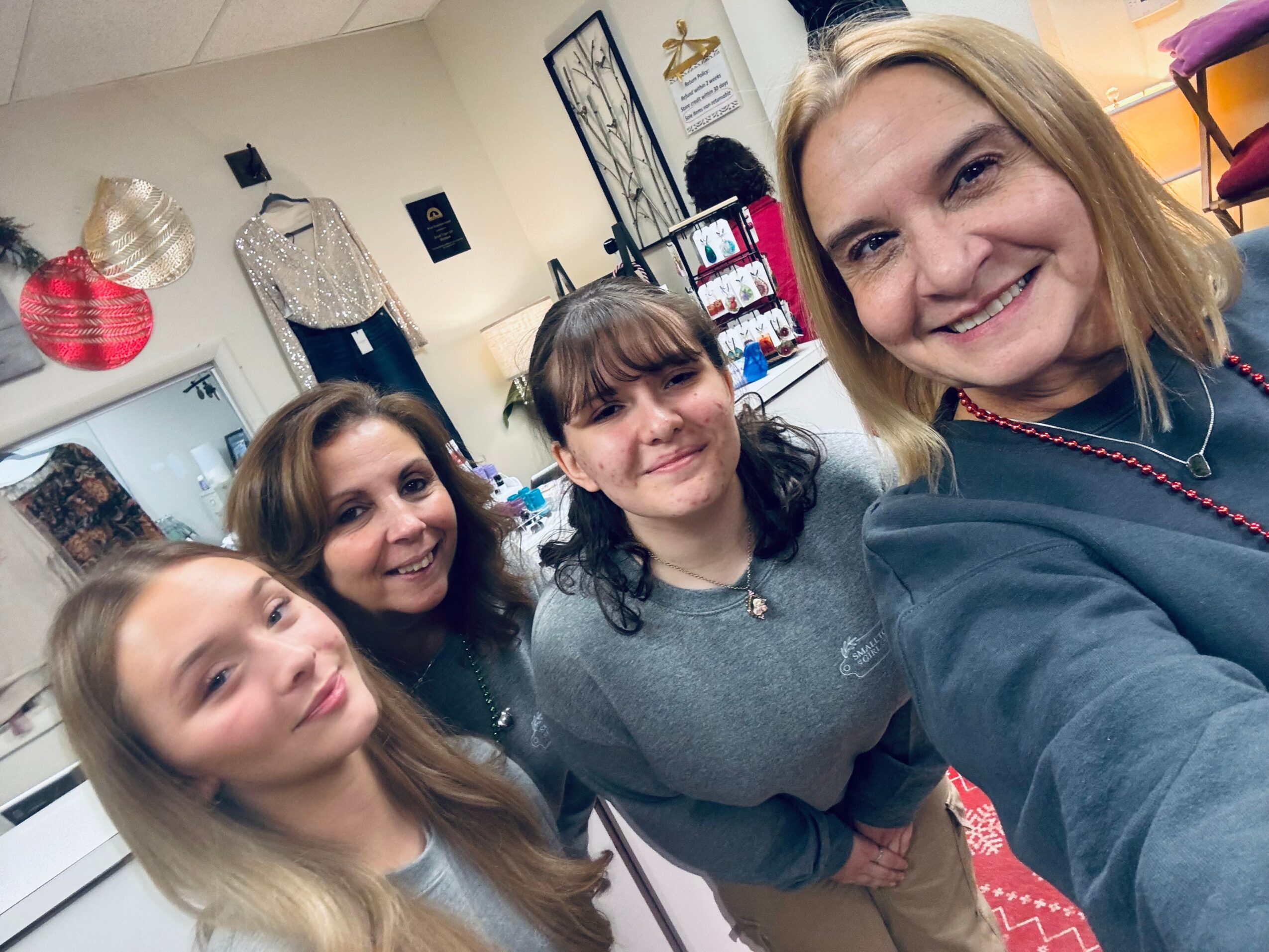 Four women smiling in a store or boutique, with clothing and accessories in the background.