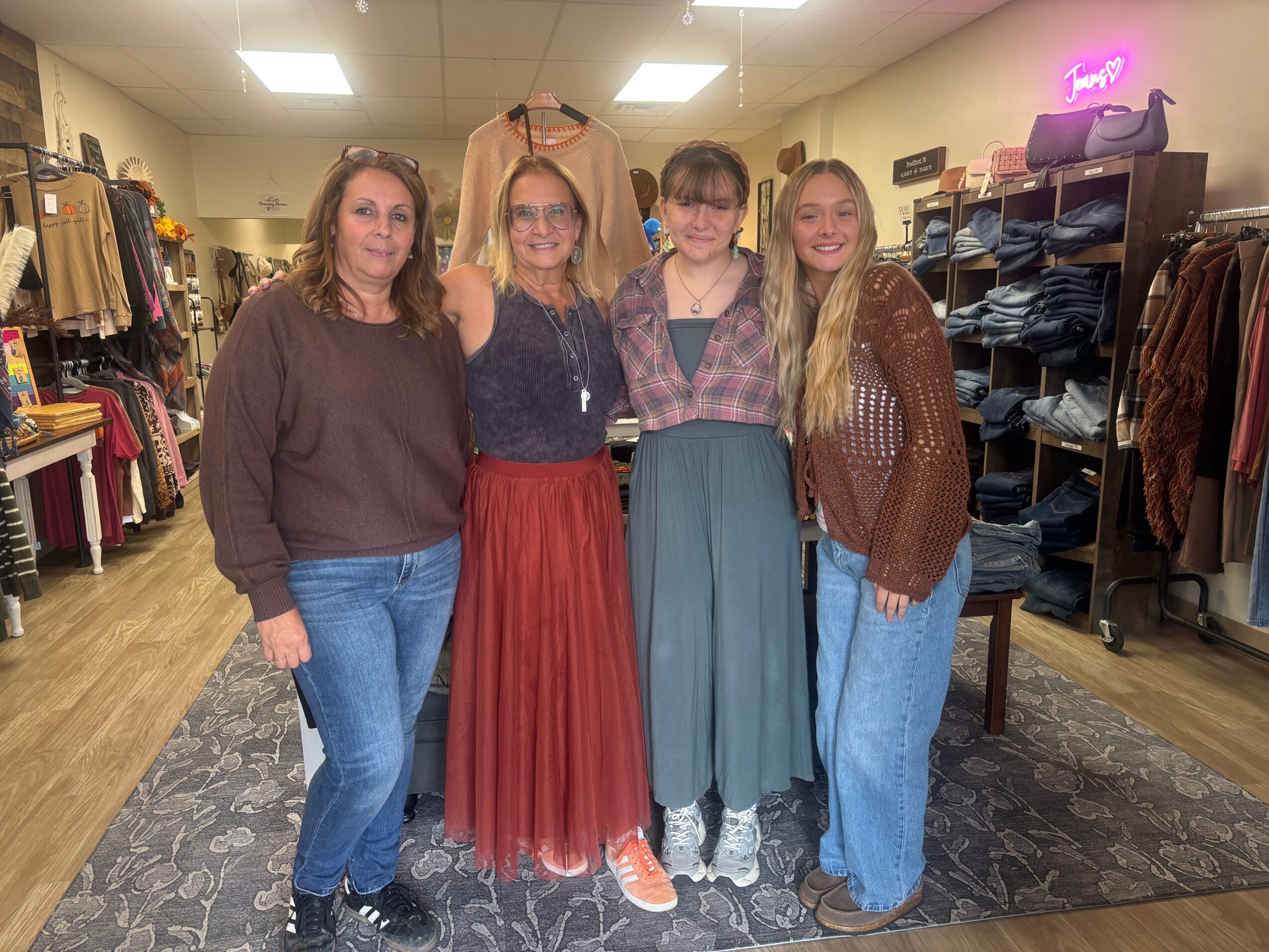 Four women standing together inside a clothing store, smiling, with racks of clothes in the background.