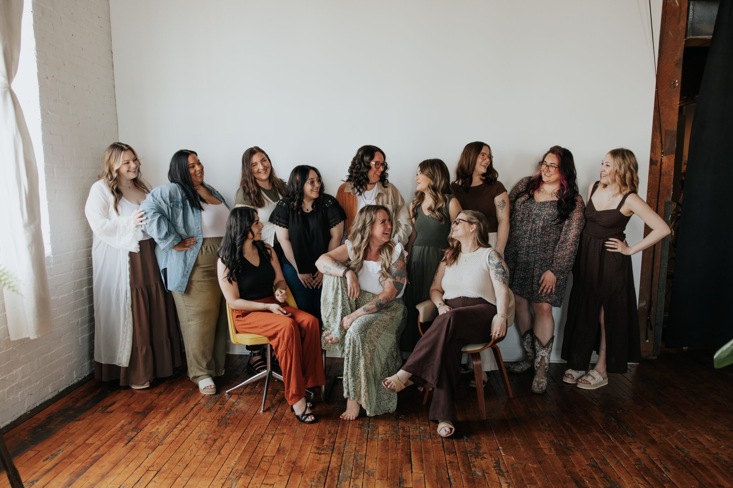 Group of women posing together indoors, some sitting and some standing, in front of a plain wall and wooden floor.