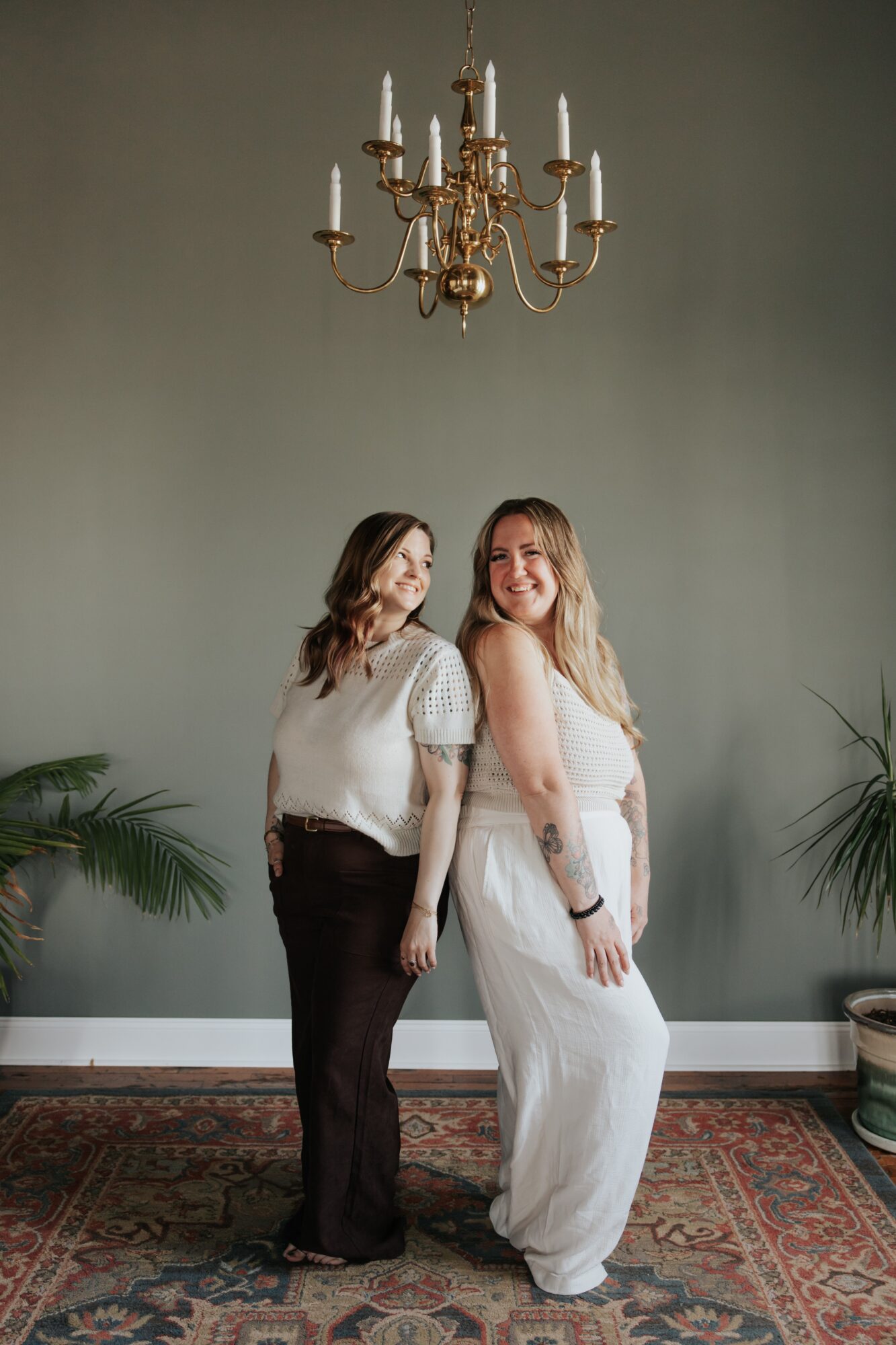 Two women stand side by side in a room with a chandelier and plants, smiling at the camera.
