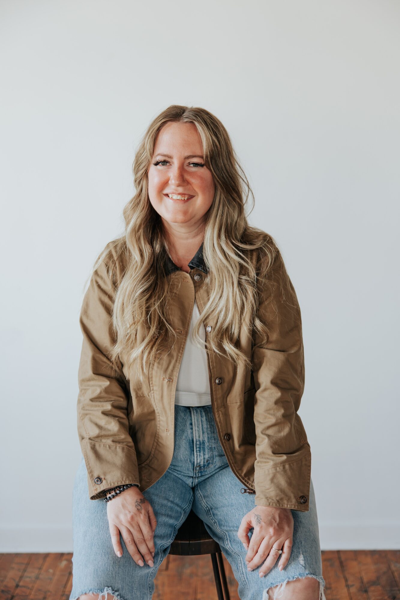 Young woman with long wavy hair smiling, wearing a brown jacket and jeans, sitting on a stool against a plain background.