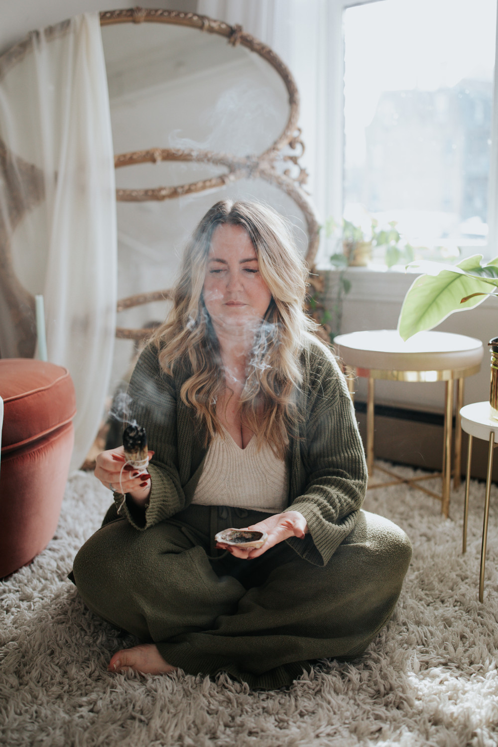Woman sitting cross-legged on a rug, holding a phone, with a plant and window in background.