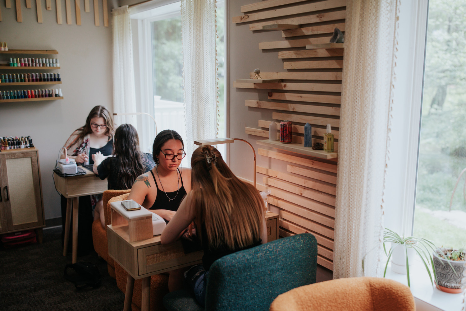 Four women sit at a table in a bright room with large windows, bookshelves, and wooden wall decor, working or studying.