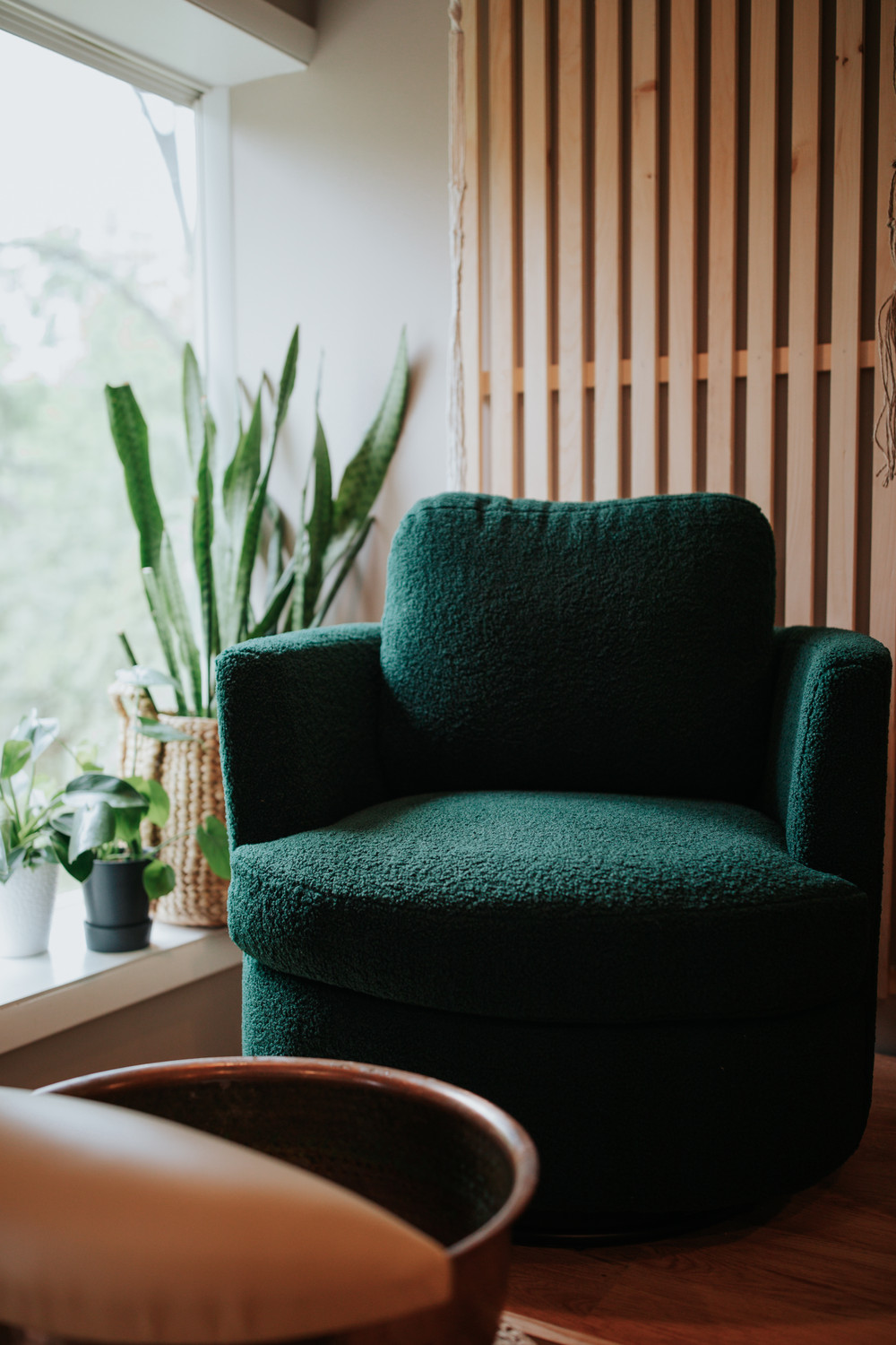 Green armchair near window with plants and wooden slat wall, natural light, cozy interior.