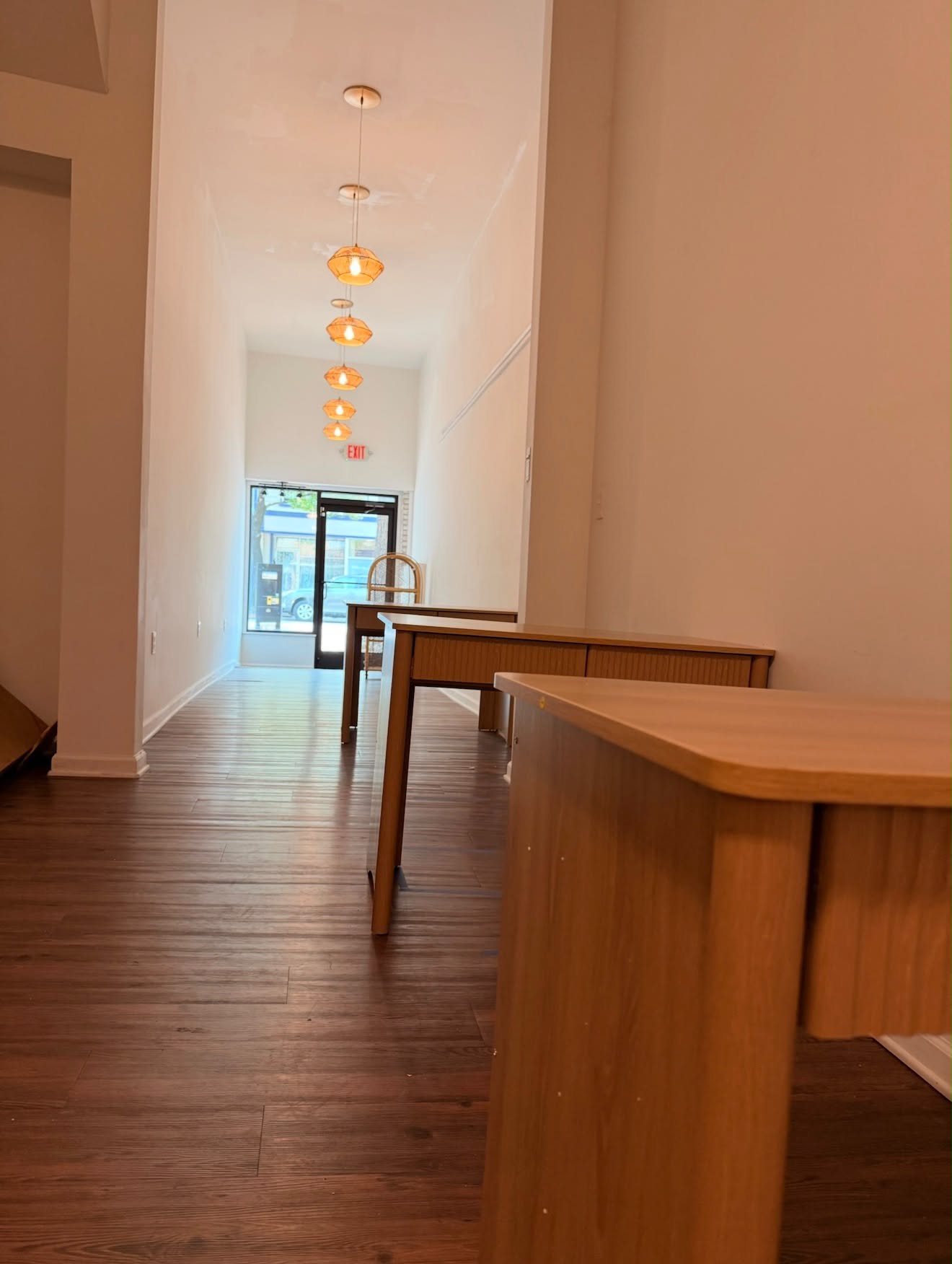 Long hallway with hanging pendant lights, wooden tables, and a door at the end, viewed from the side.