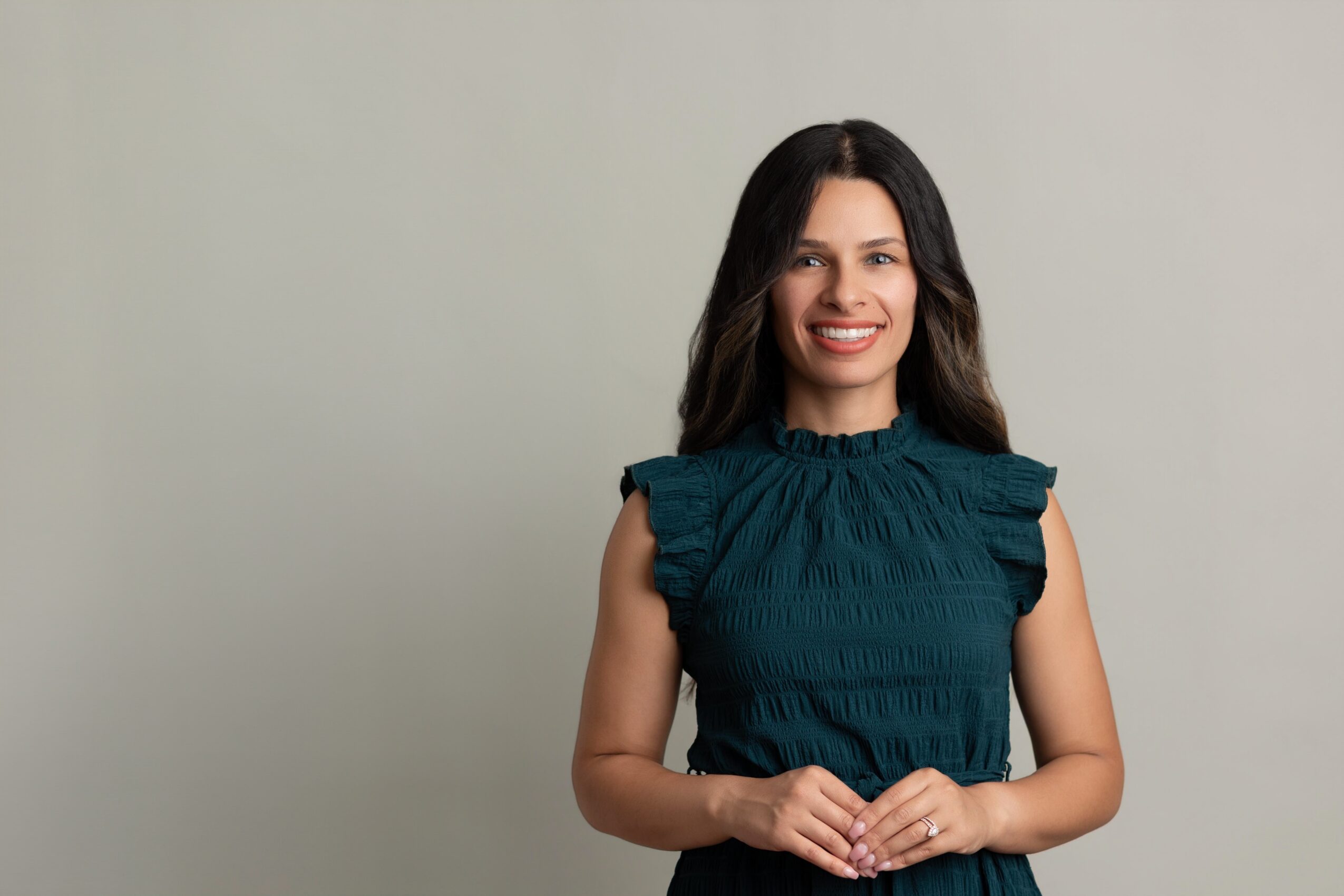 Young woman with long dark hair smiling, wearing a teal sleeveless top, standing against a plain light background.