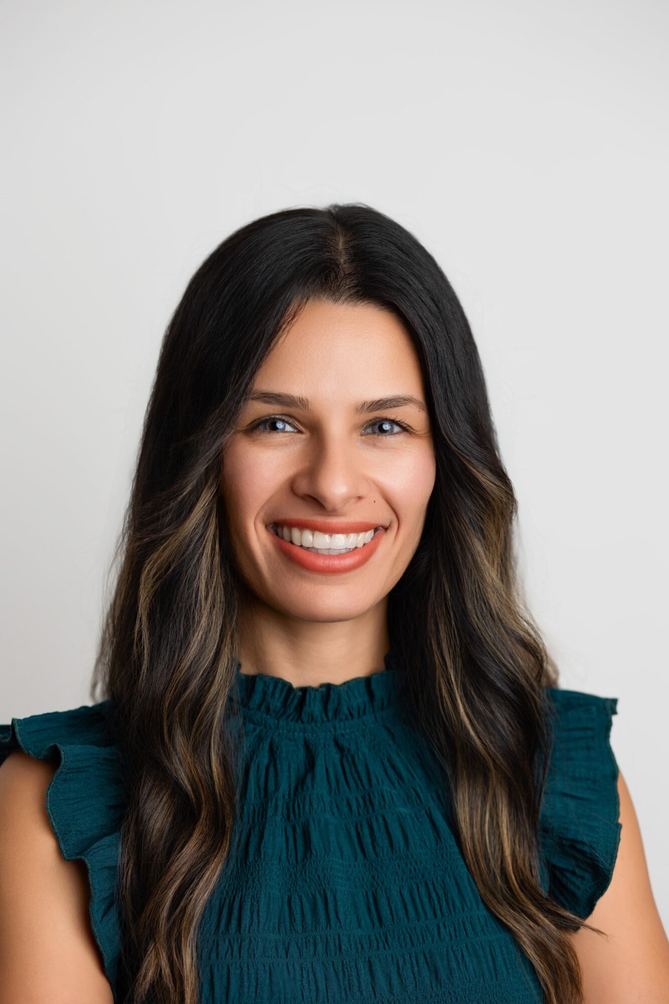 Smiling woman with long dark hair wearing a teal top against a plain background.