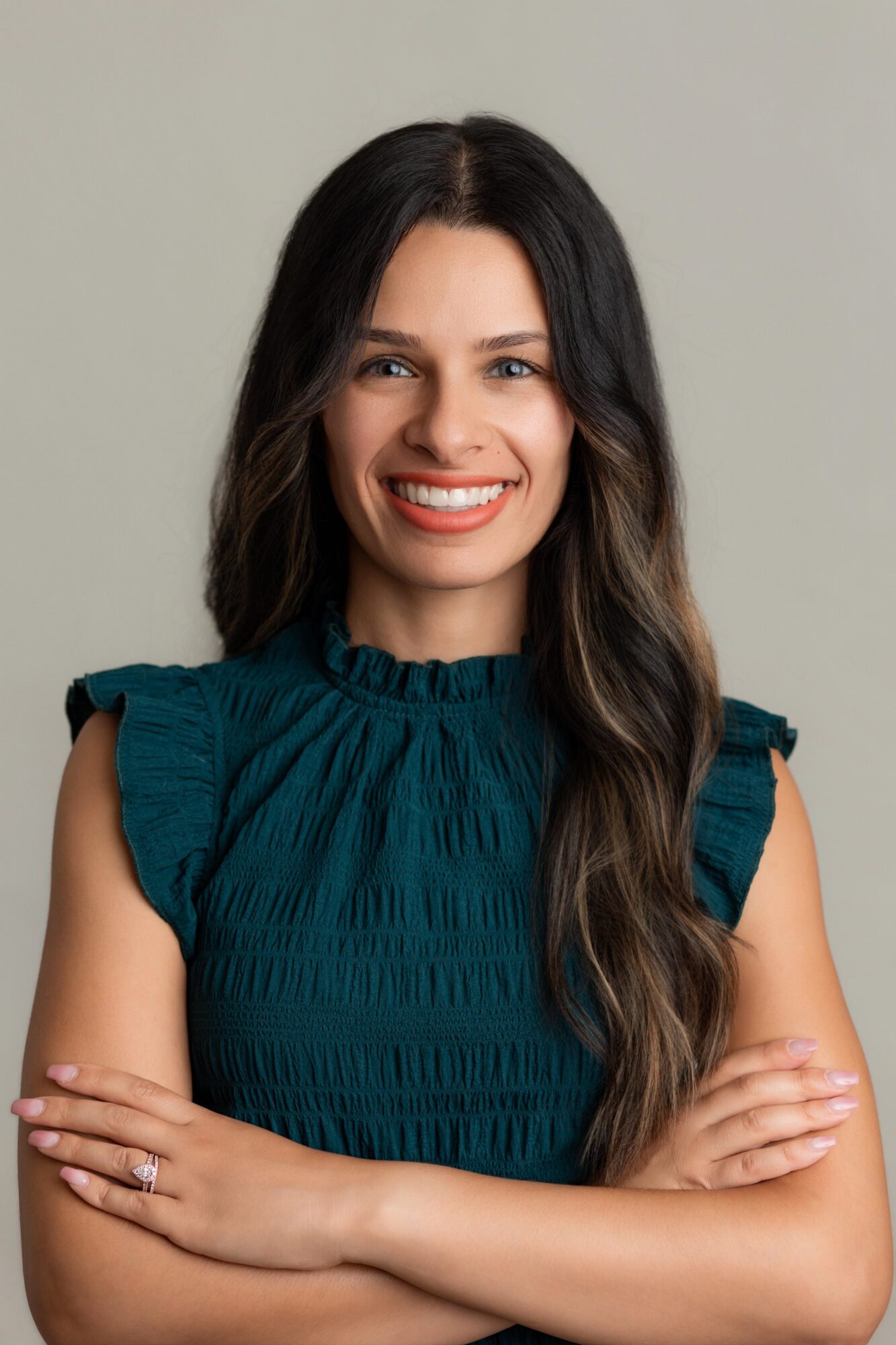 Smiling woman with long dark hair wearing a teal ruffled top, arms crossed, against a plain background.