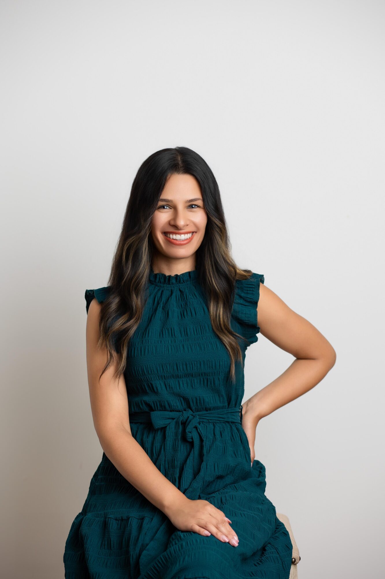 Young woman with long dark hair smiling, wearing a teal dress, standing against a plain white background.