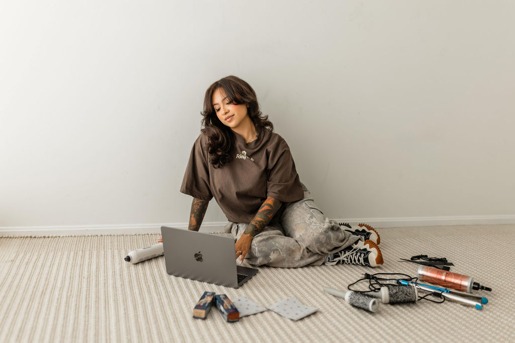 Young woman sitting on a carpeted floor with a laptop, surrounded by various tools and electronic components, against a plain wall.