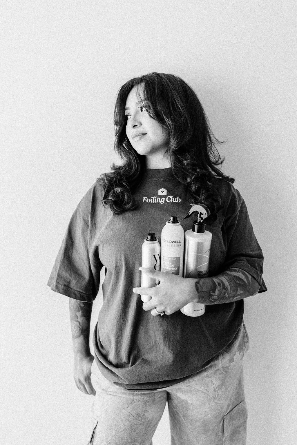 Woman with dark hair holding bottles, wearing a loose T-shirt, standing against a plain wall.