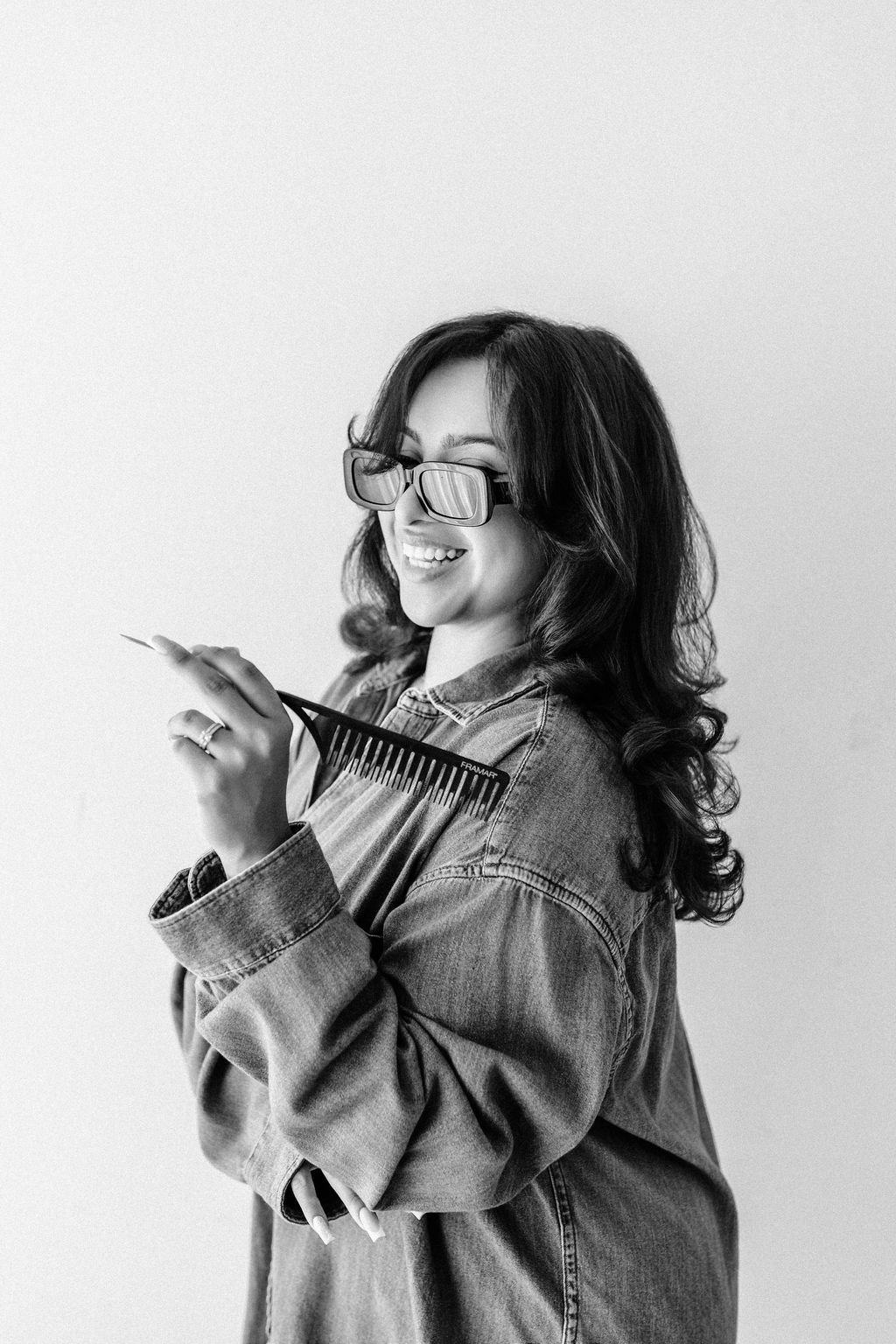 Smiling woman with glasses and long hair, holding a comb and a hairbrush, wearing a denim jacket, in black and white.
