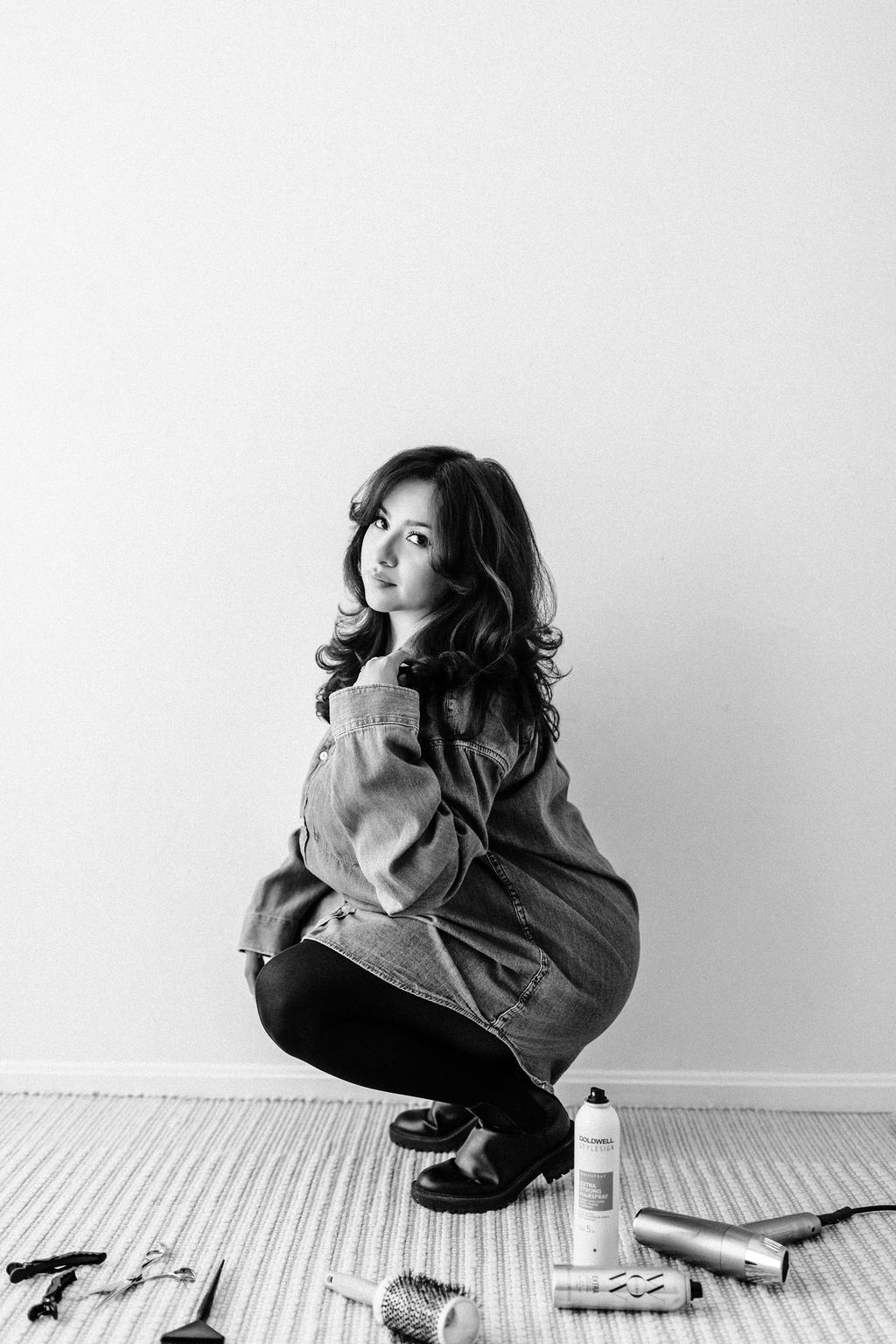 A woman crouching on a striped rug, with hair styled in loose waves, wearing a denim jacket and black shoes, against a plain wall.