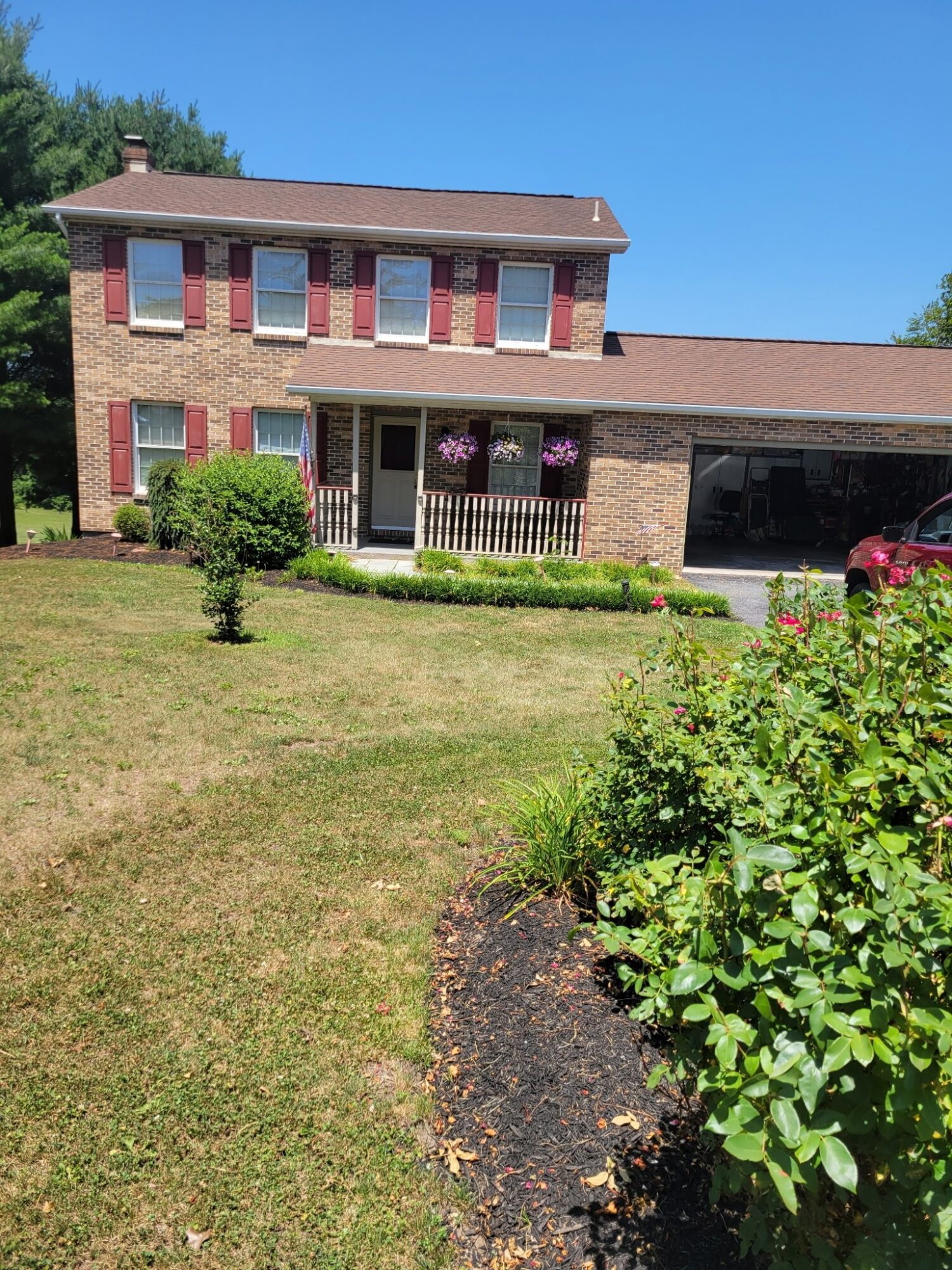 Two-story brick house with a front porch, surrounded by a lawn, bushes, and a garden bed, under a clear blue sky.