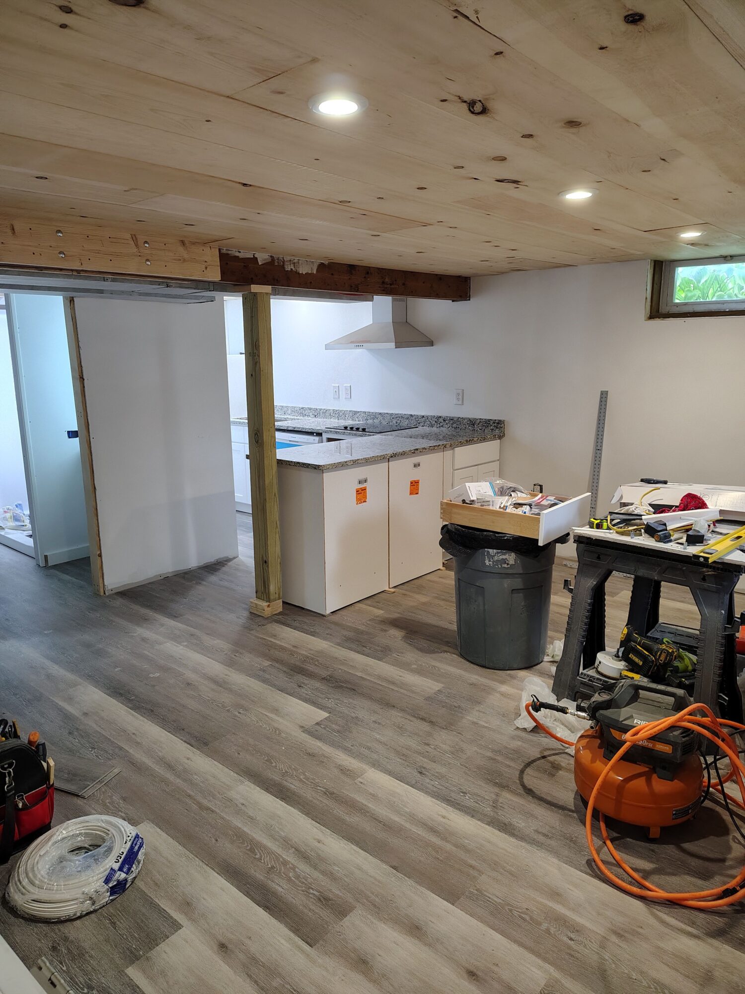 Room with wooden and laminate flooring, partially finished ceiling, and construction tools and materials visible.