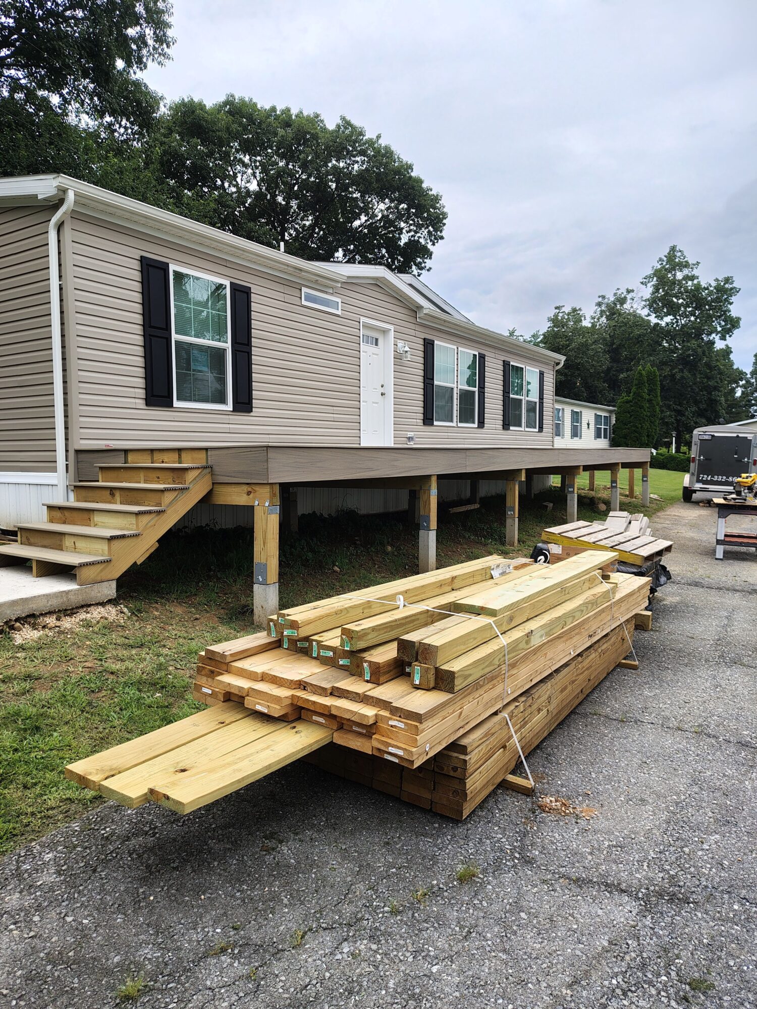 Stack of wooden planks and beams outside a house with beige siding, on a gravel driveway.