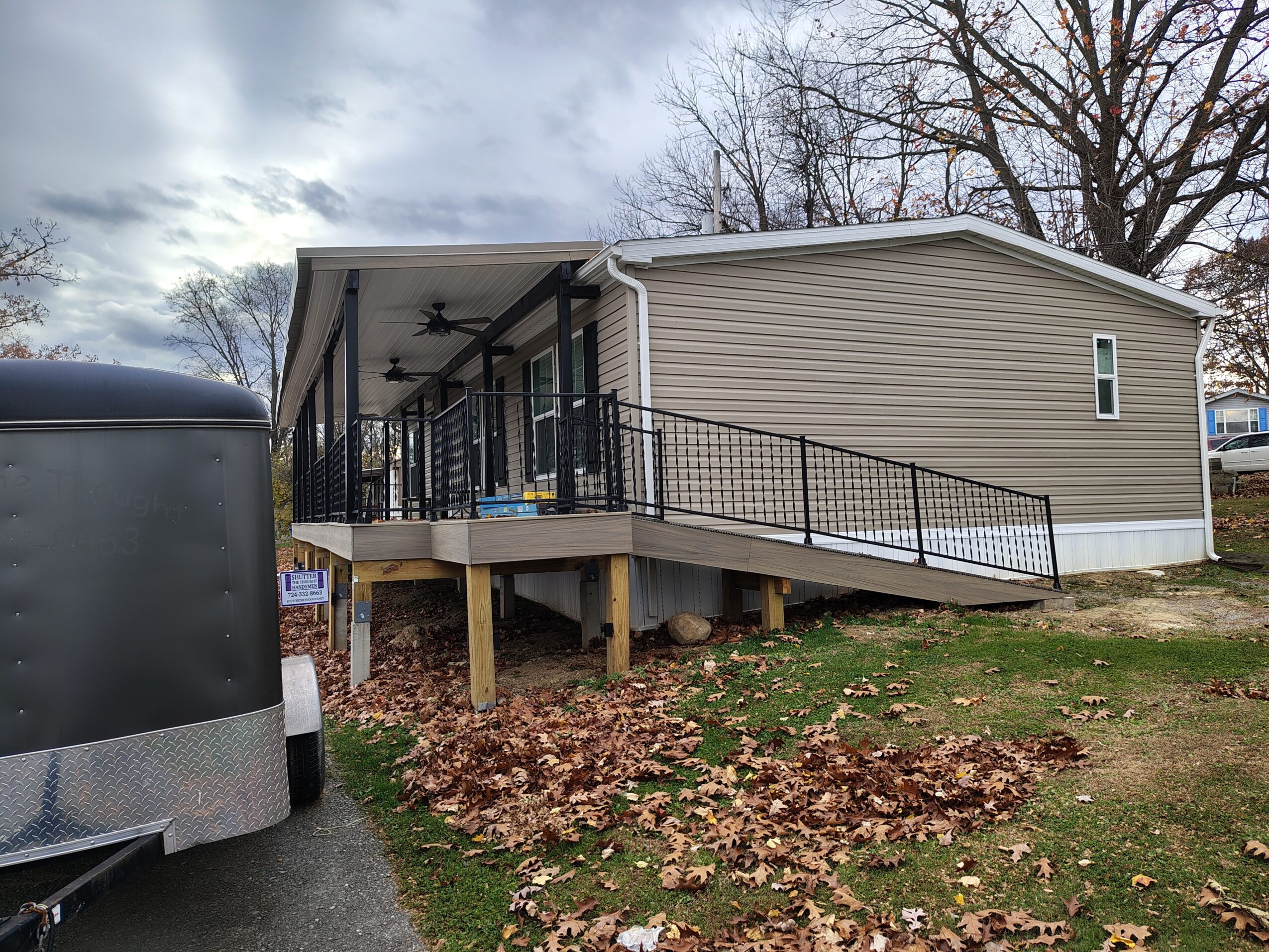 Small beige building with a ramp, black railing, and a covered porch, elevated on wooden supports, with trees and cloudy sky in background.